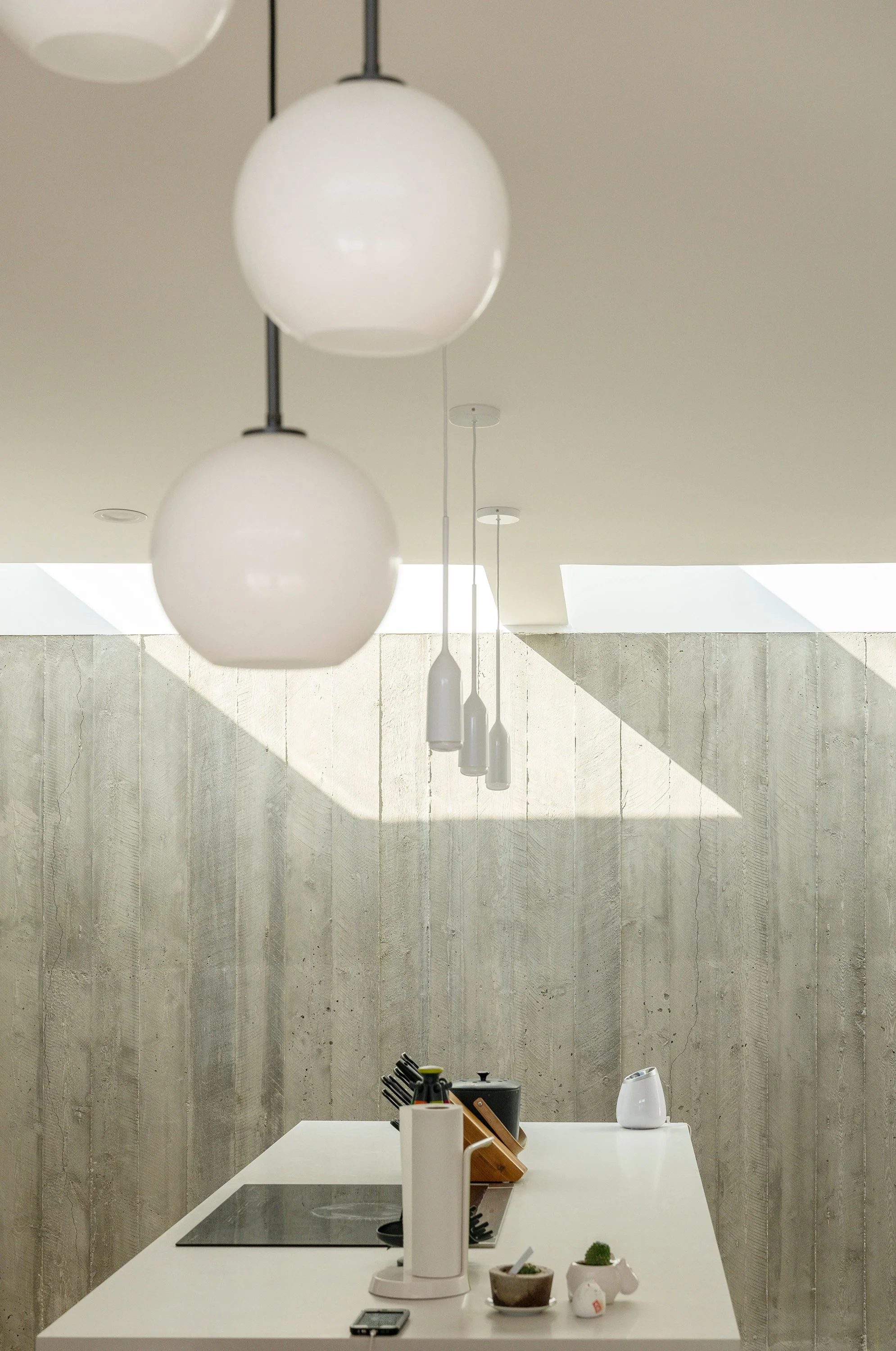 A kitchen island with an induction cooktop and pendant lights is illuminated by natural light through a square skylight above a textured concrete wall.