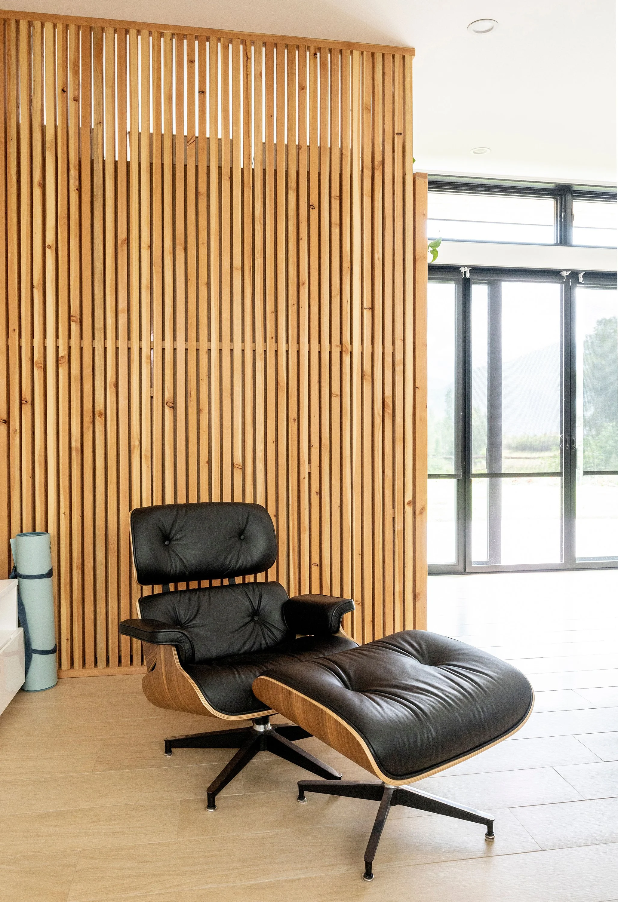 Modern lounge chair and ottoman placed near vertical wood slat partition and large windows with black frames