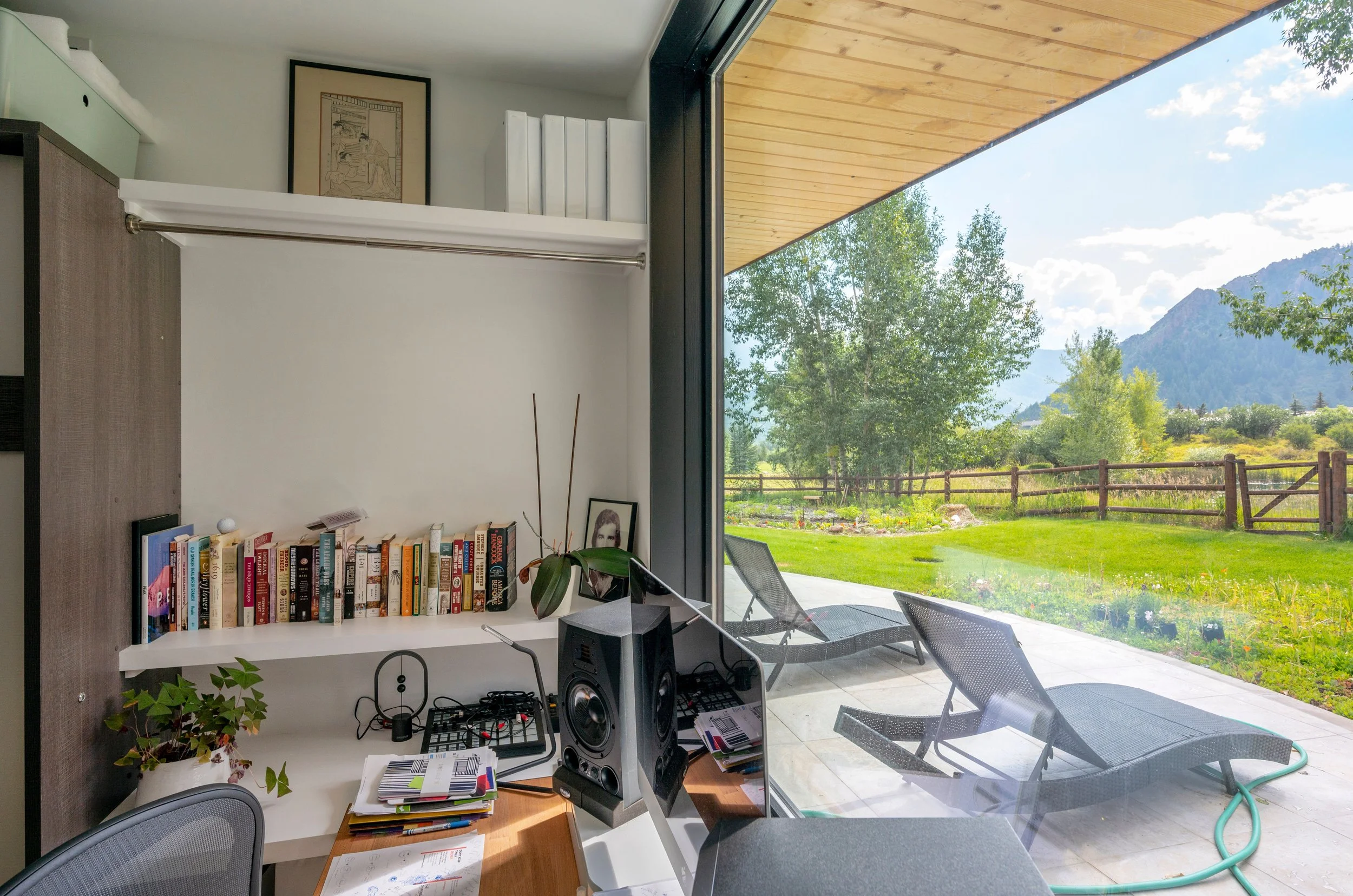 A home office space with a large picture window opening to a patio featuring lounge chairs and a fenced garden area.