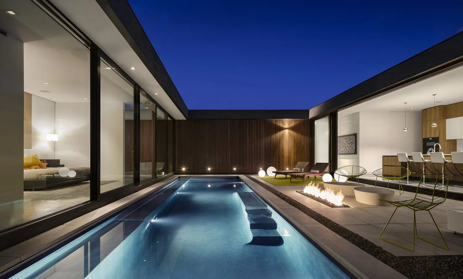 Modern courtyard with pool and fire pit between living and kitchen areas at night.
