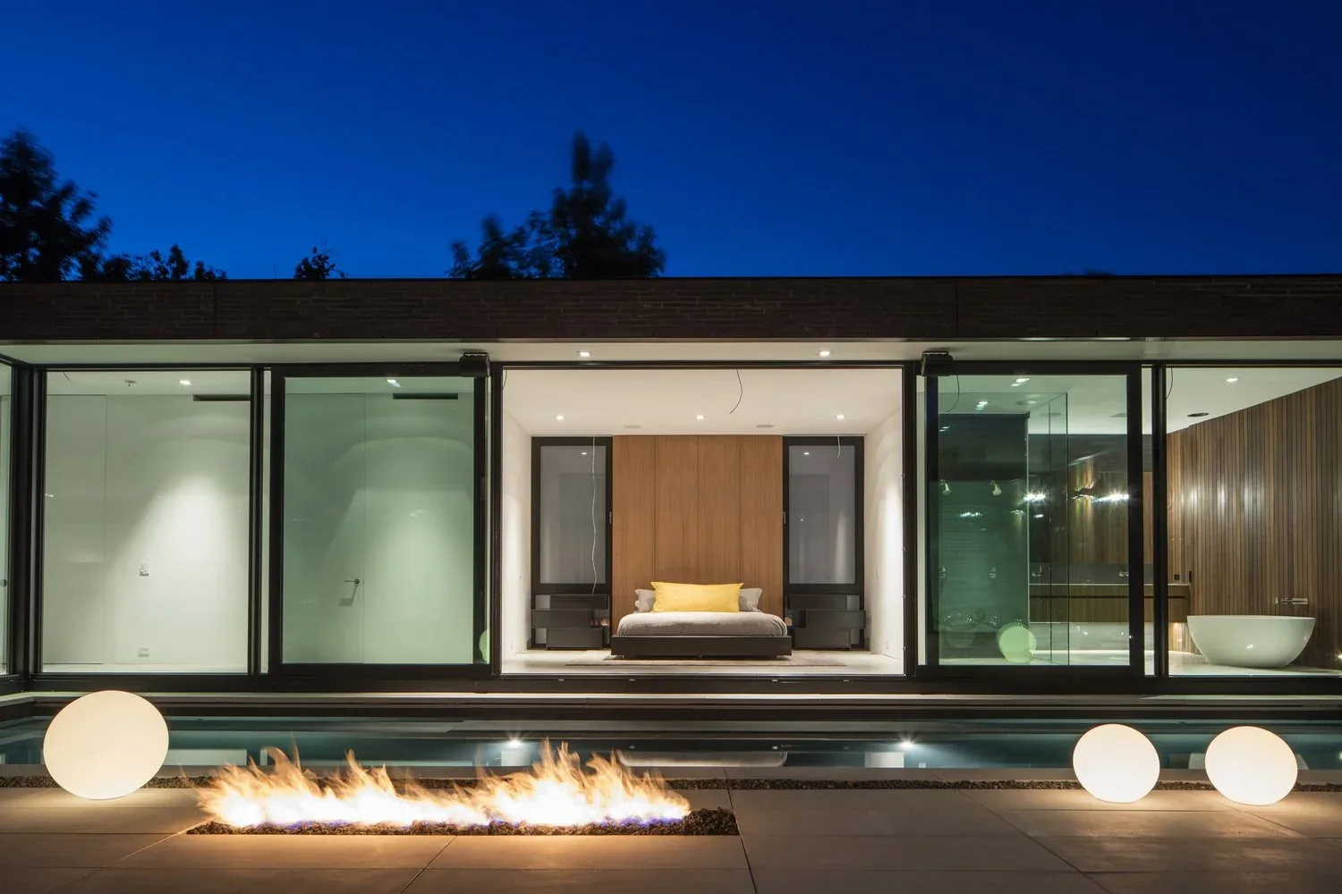 The exterior rear view shows a bedroom and bathroom with floor-to-ceiling glazing, a fire pit, and a pool at night.