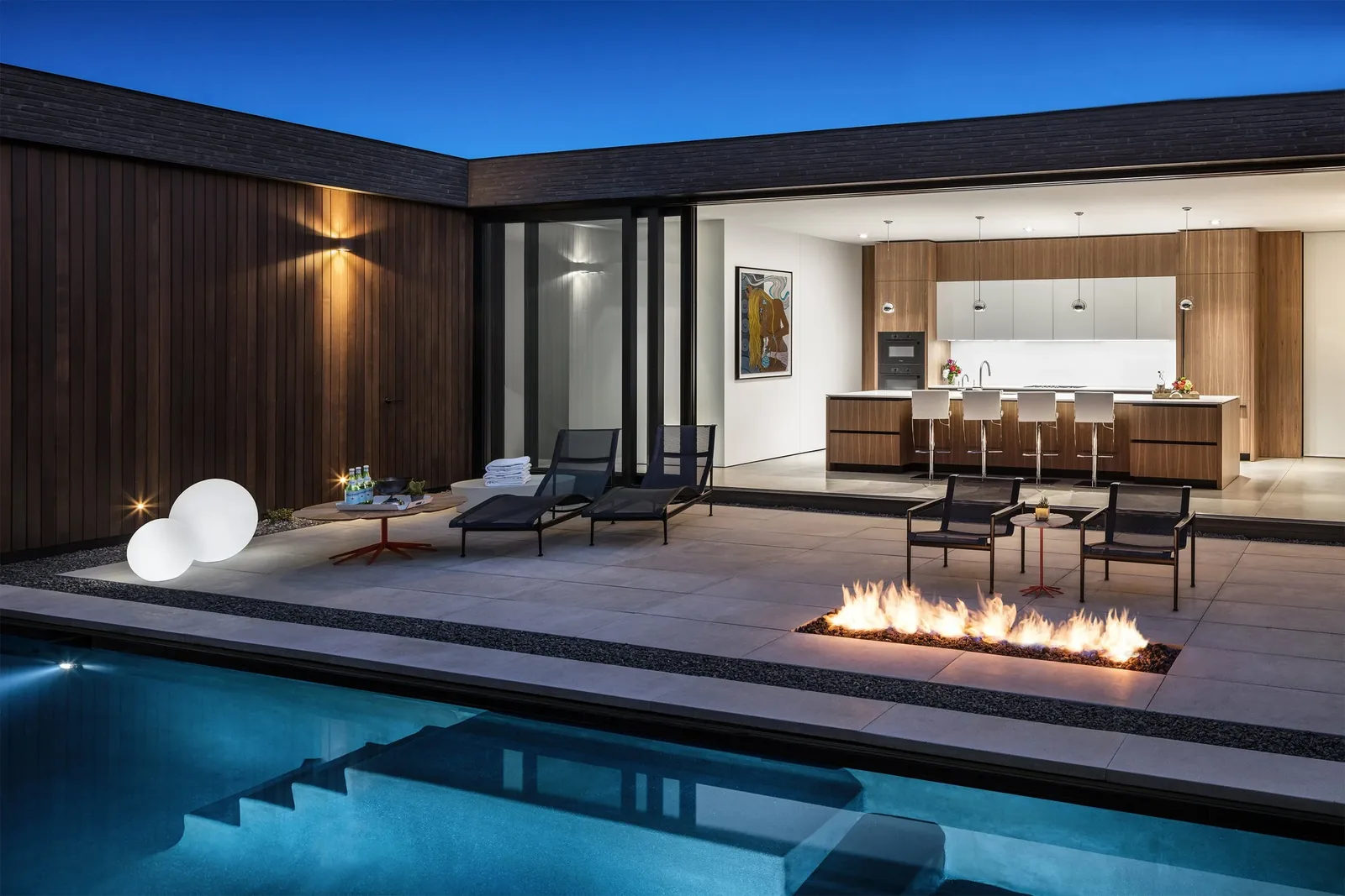 View of patio with fire pit and pool fronting an open kitchen in a contemporary house at dusk.