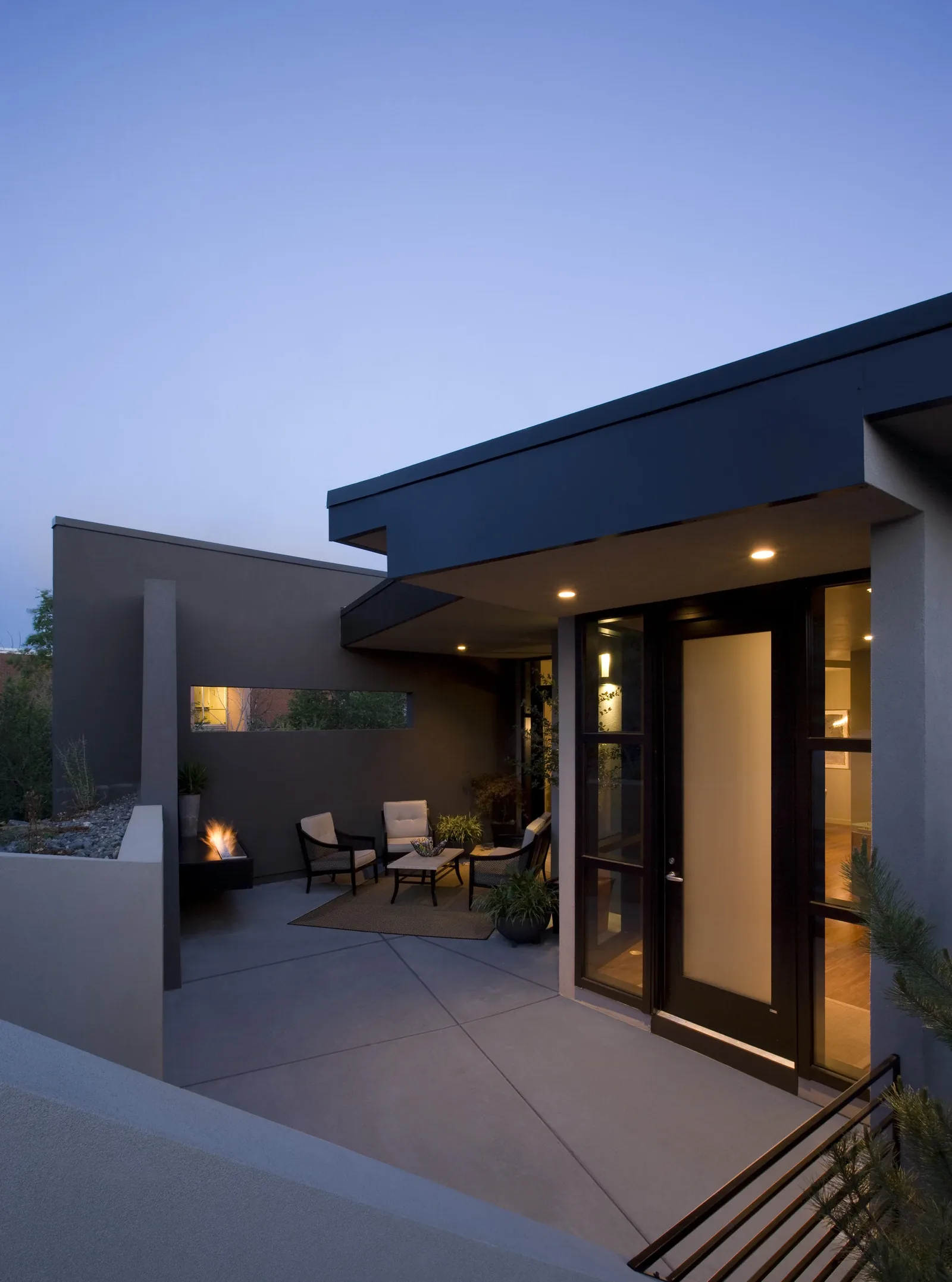 Covered outdoor seating area and the front entry of the modern Fractured Residence at dusk.
