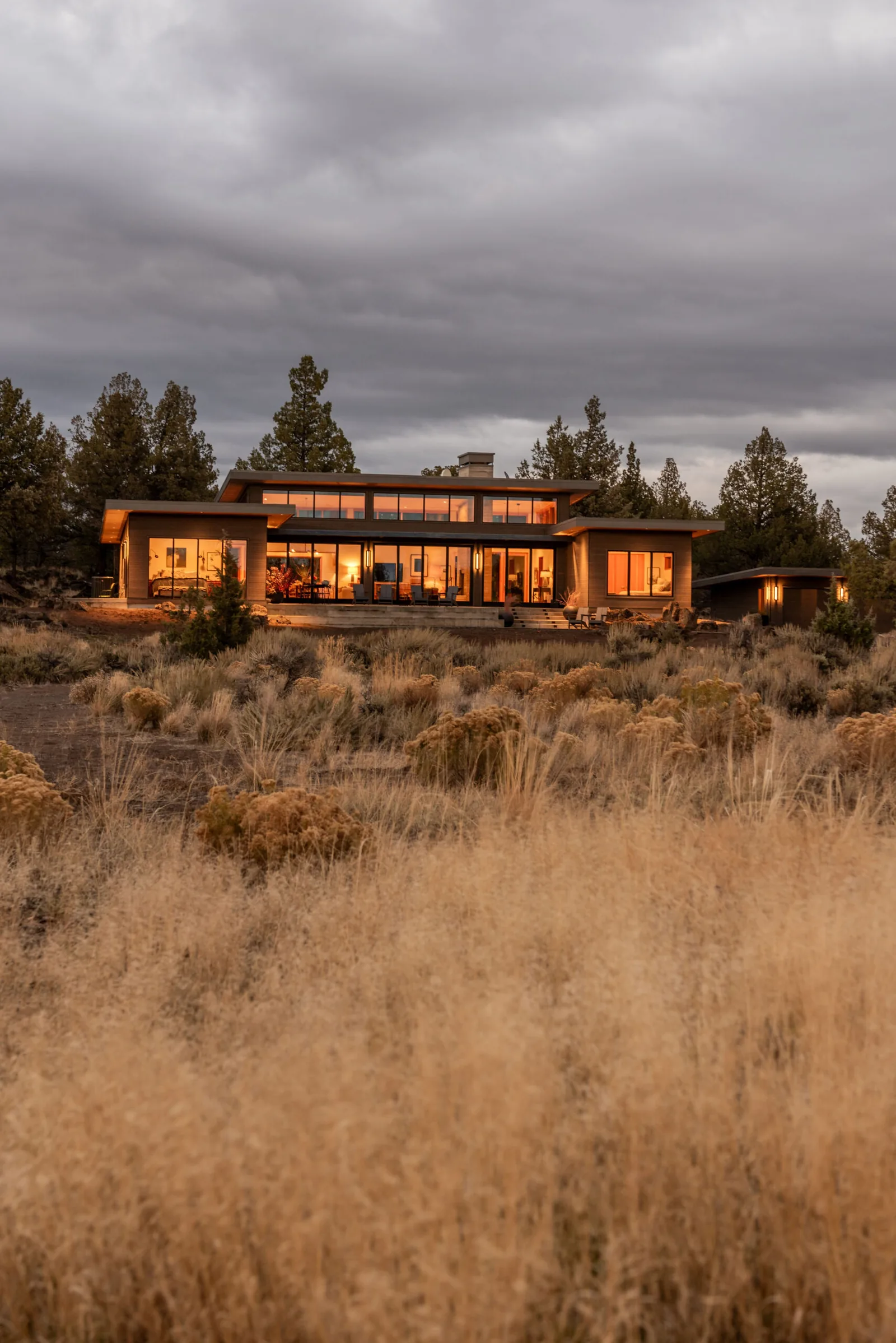 Exterior front view of a modern home illuminated at dusk, set in a natural landscape with trees and dry grass.