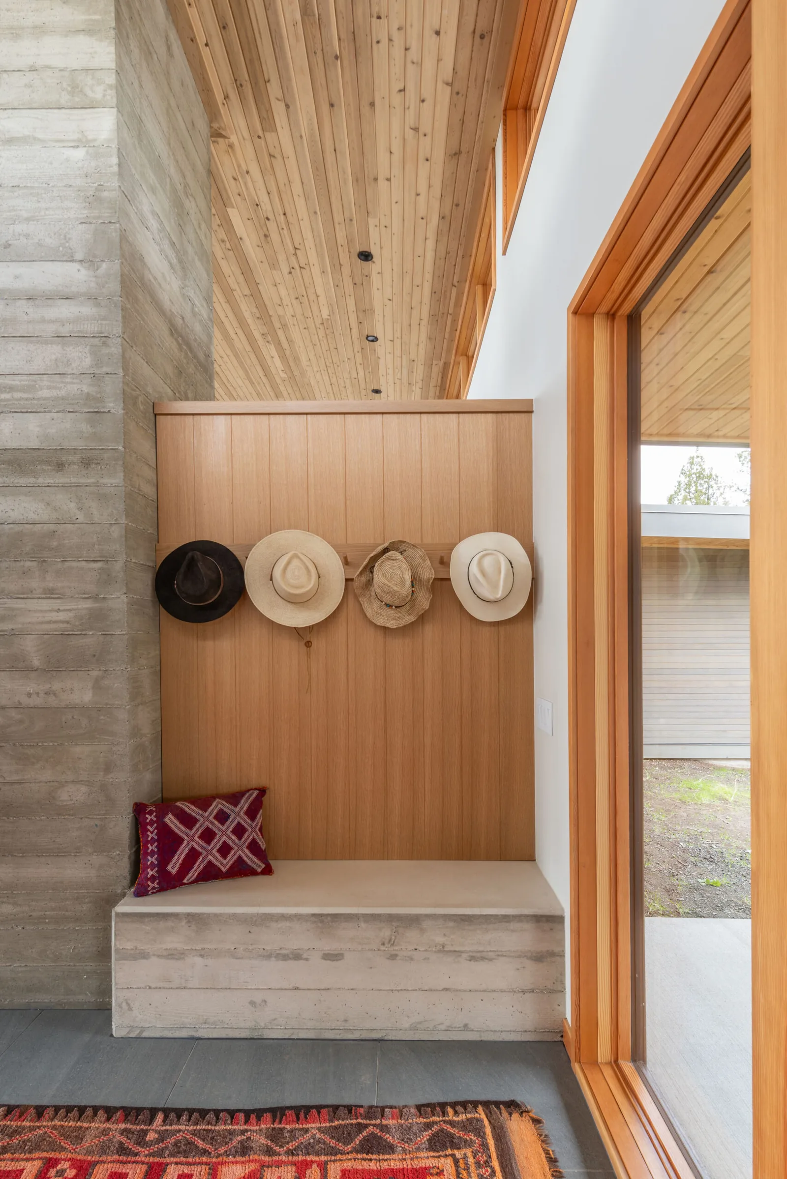 Entry with concrete bench, wood panel wall, hat hooks, and views to the exterior through large wood-framed glass door.