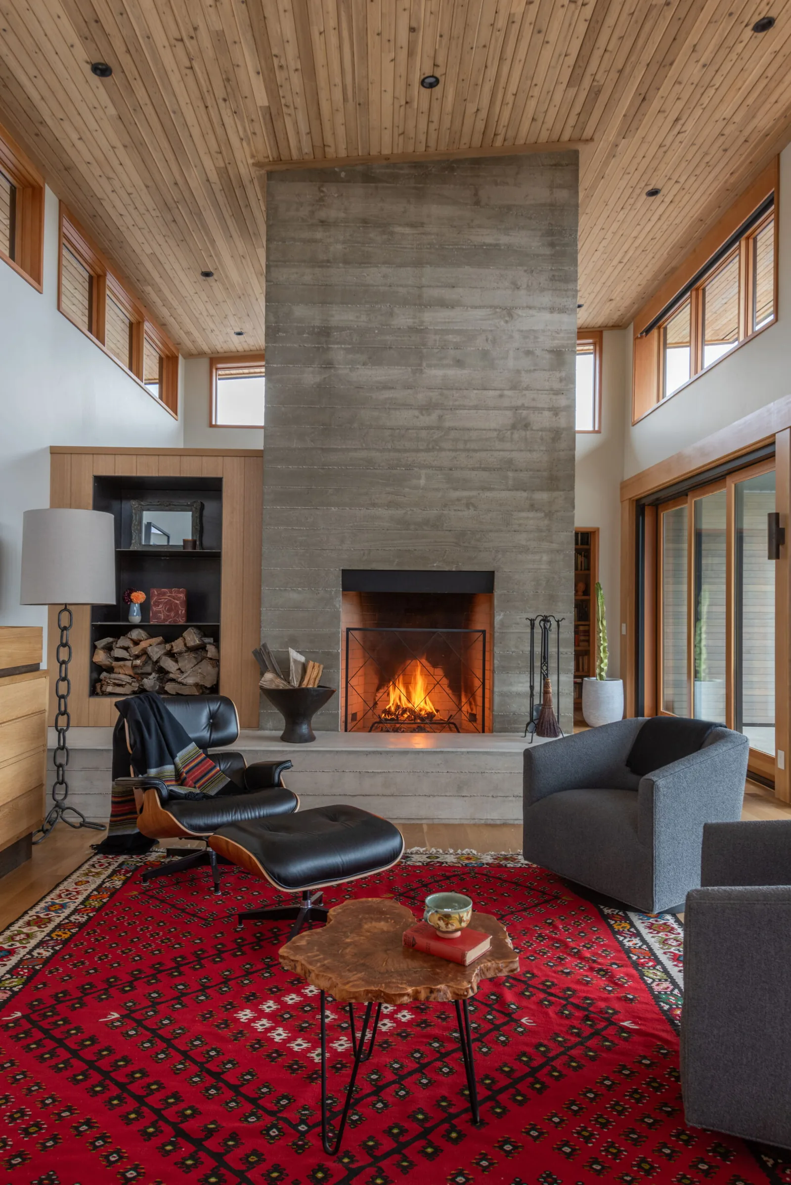 Living room features a tall concrete fireplace, wood ceiling, clerestory windows, and sliding glass doors, blending modern and natural materials.