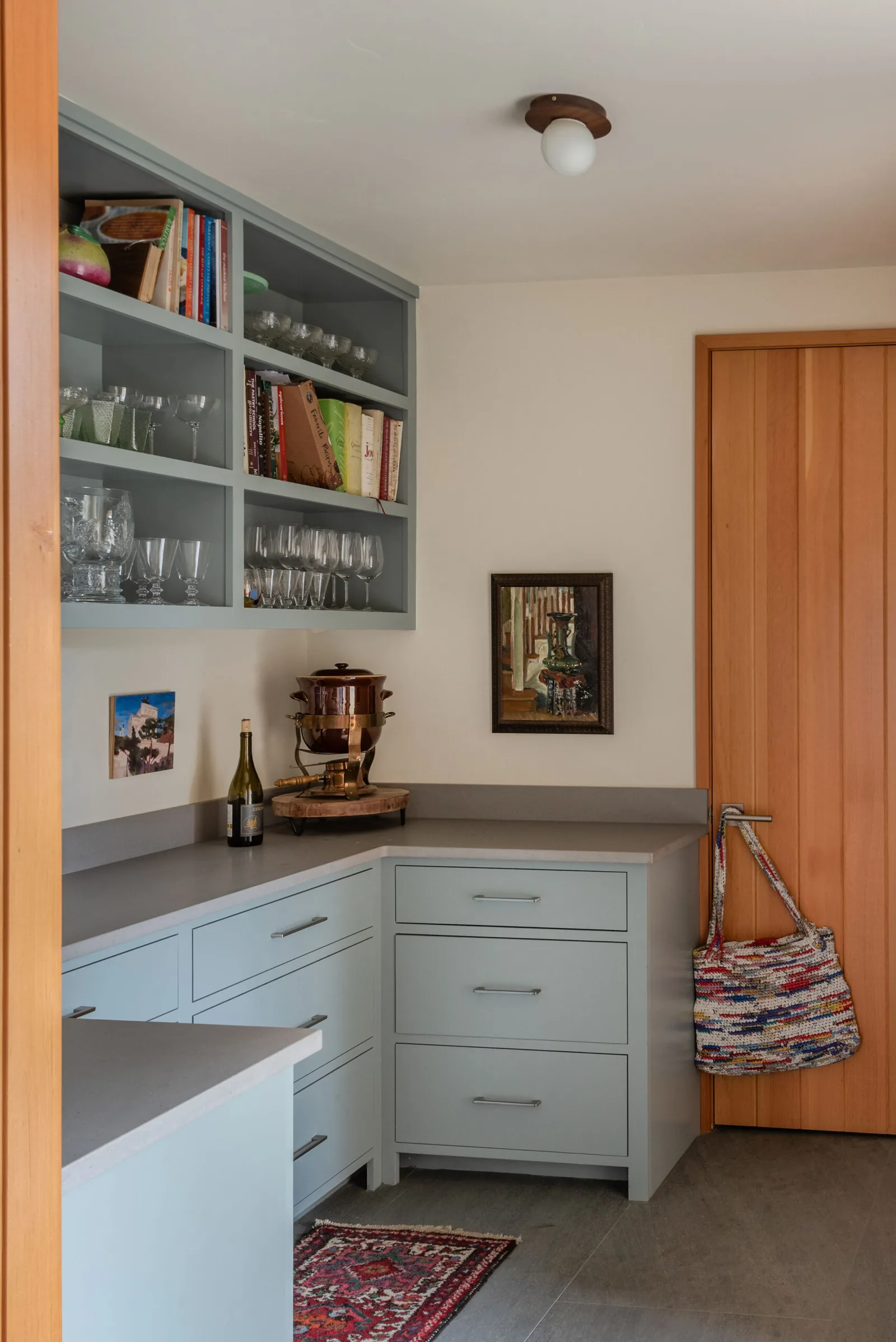 Light blue kitchen cabinets and open shelves with glassware and books, adjacent to a wooden door with a woven bag.