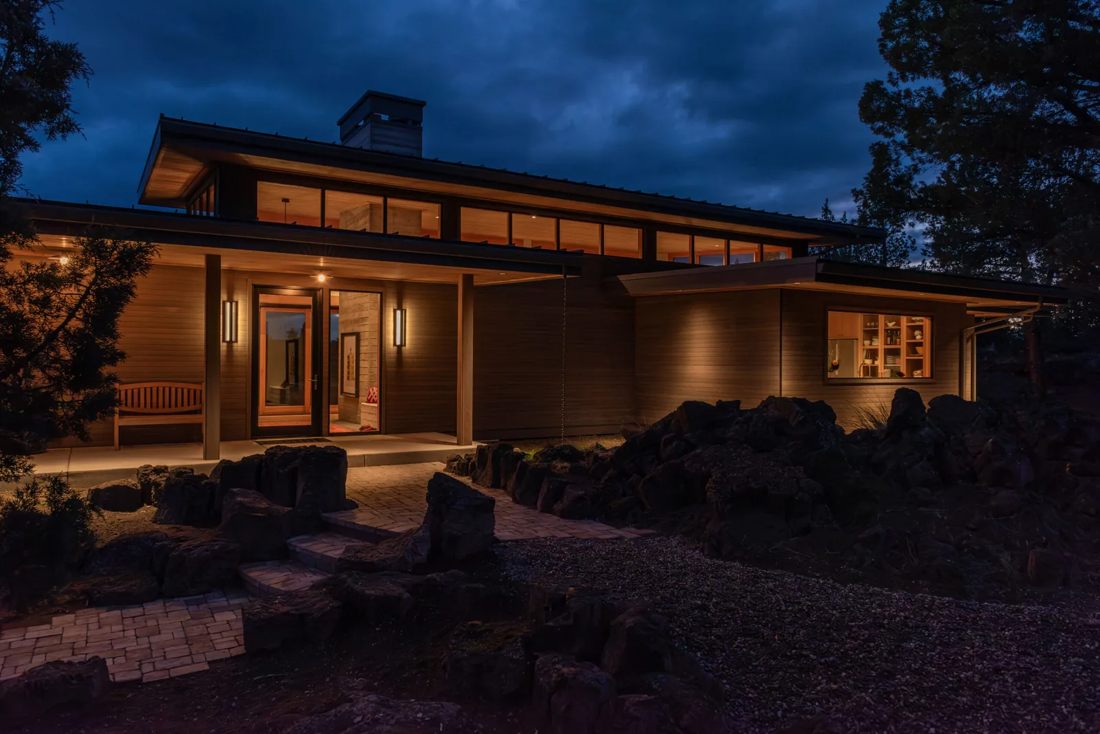 Modern style house entrance illuminated at dusk with rock landscaping and a bench seating area by the door.