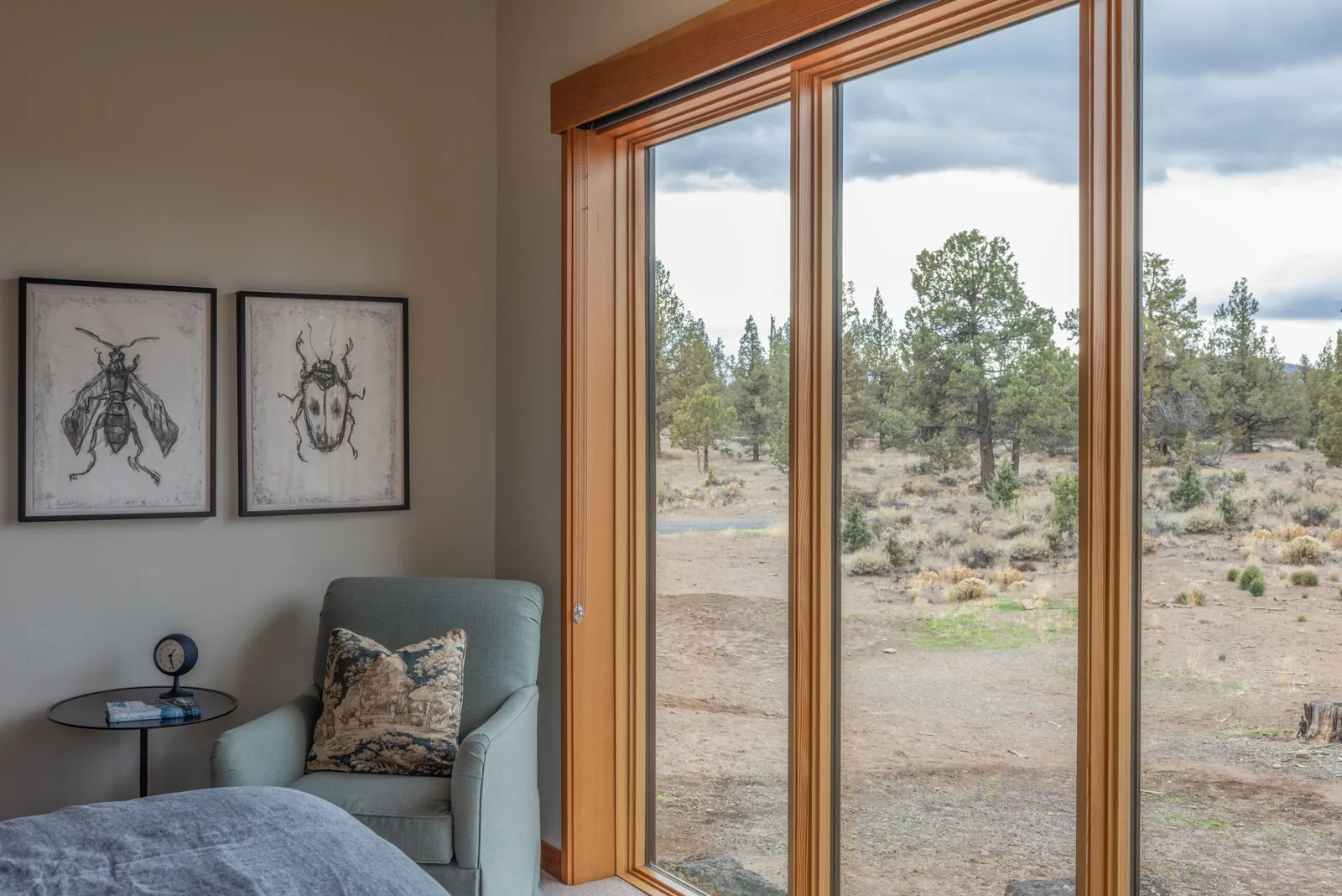 Bedroom corner with light blue armchair and large wooden framed windows overlooking desert landscape in DESCHUTES MODERN OASIS.