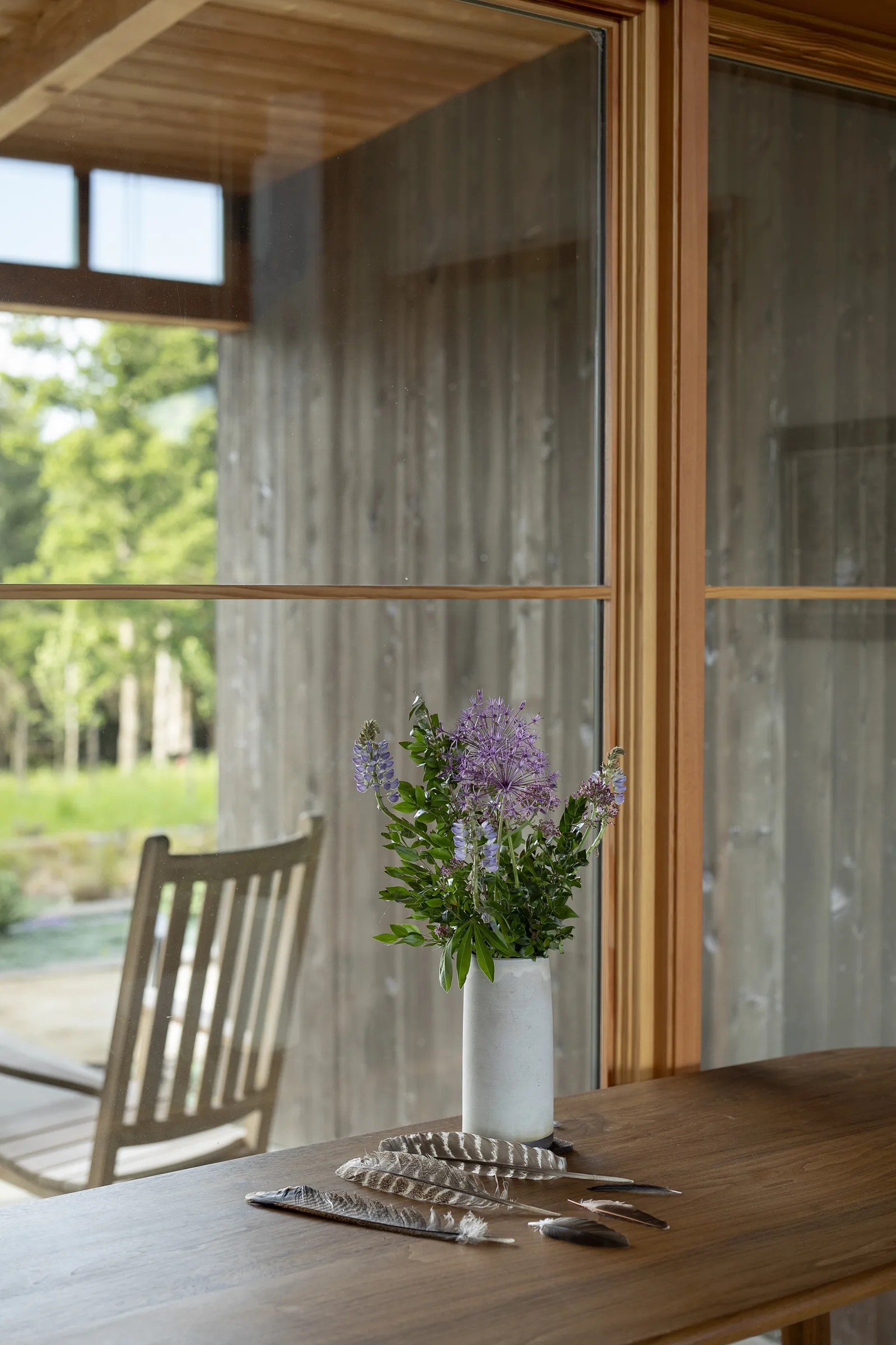 Interior view of a wooden table with vase and feathers in front of a large wood-framed window overlooking a patio with outdoor chair.