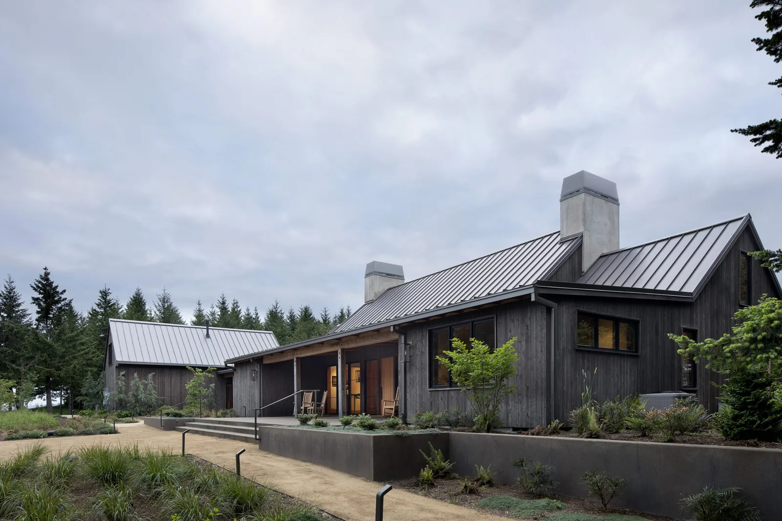Exterior front view of a wood-clad farmhouse with metal roofs and concrete chimneys at dusk.