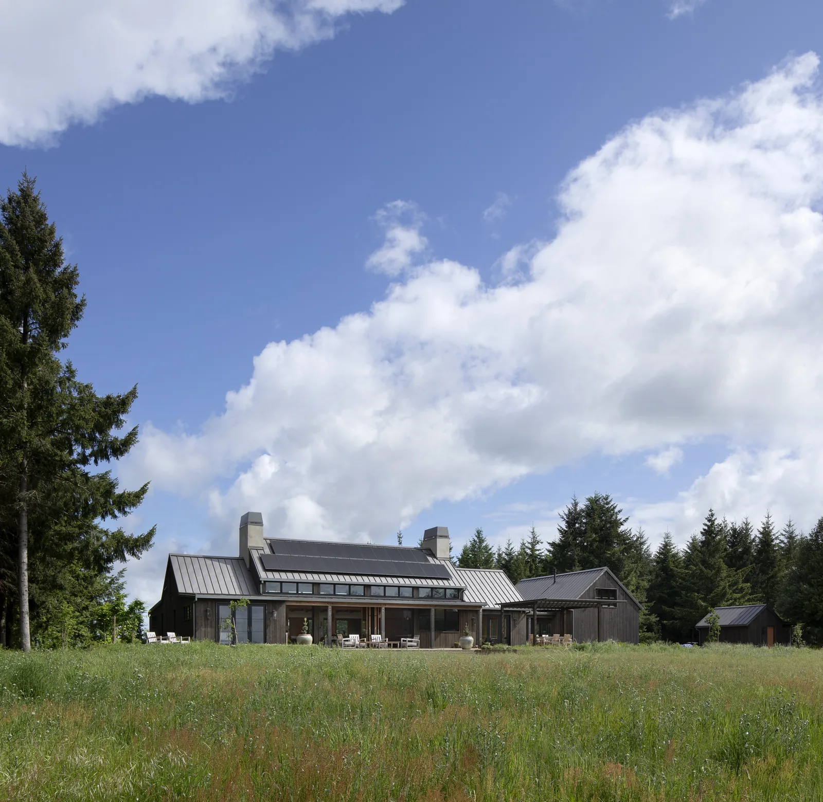 Dark wood farmhouse with metal gable roofs and solar panels in a rural field setting.