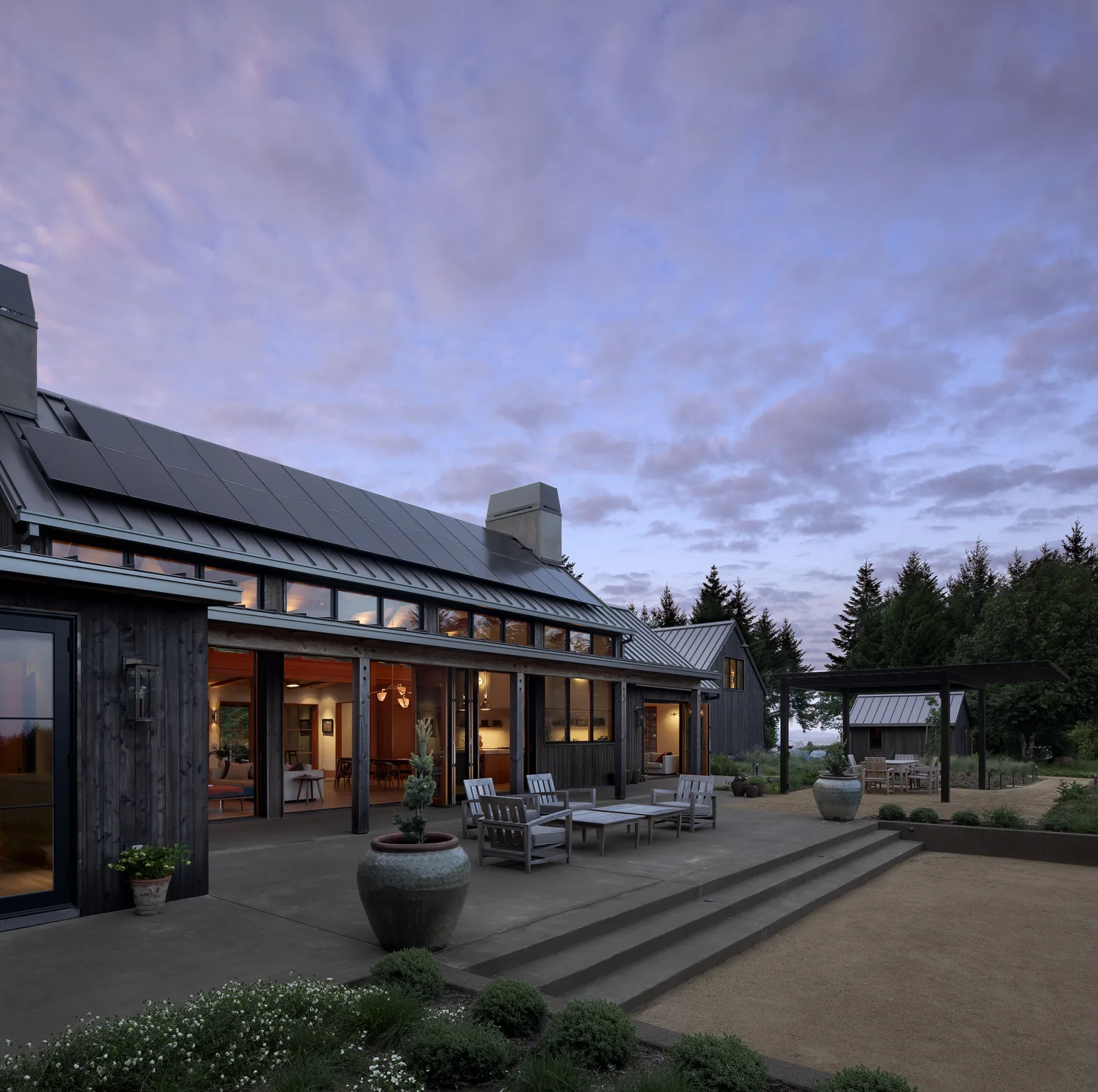 Exterior wide view of a modern house with a metal roof, sliding glass doors, and an outdoor patio seating area.
