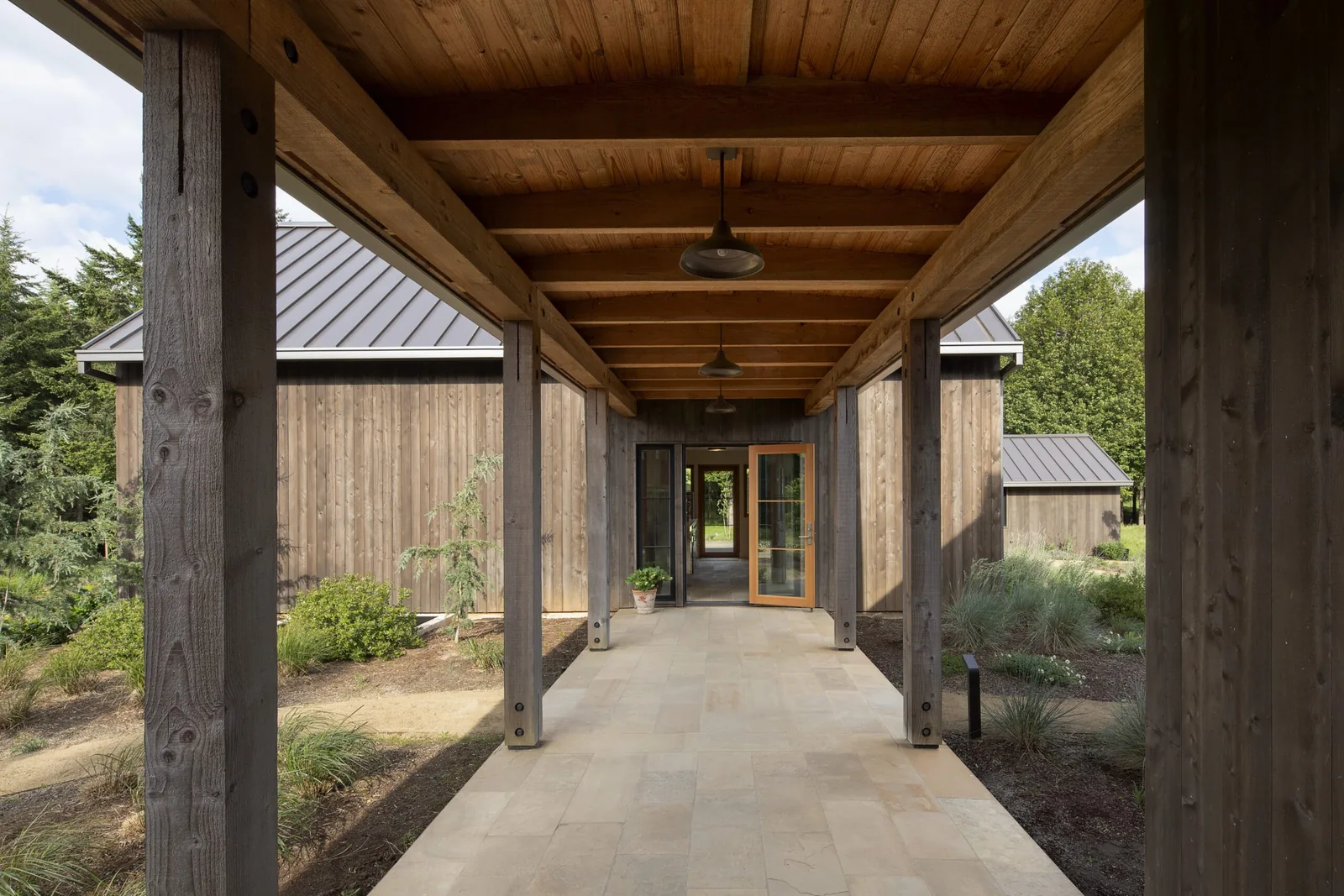 View through a covered exterior corridor with exposed wooden beams, leading to a glass and wood entry door.