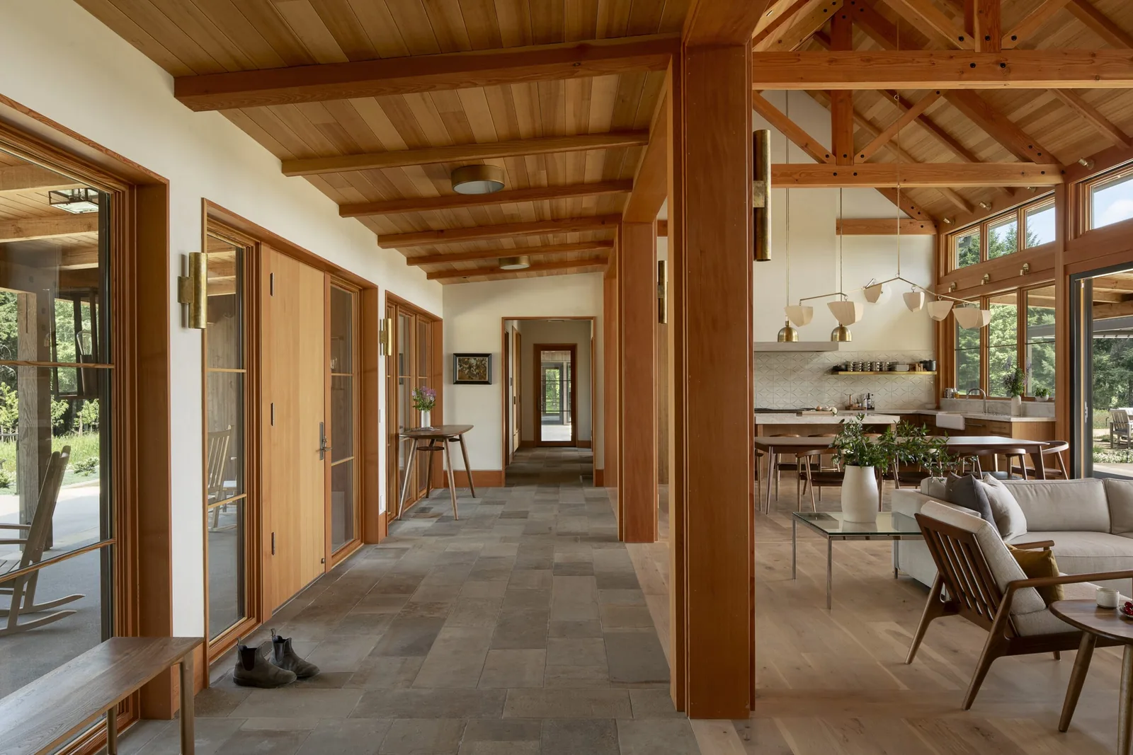 Interior hallway with wood beams and stone tile floor opens into a living area with vaulted wood ceiling and large windows.