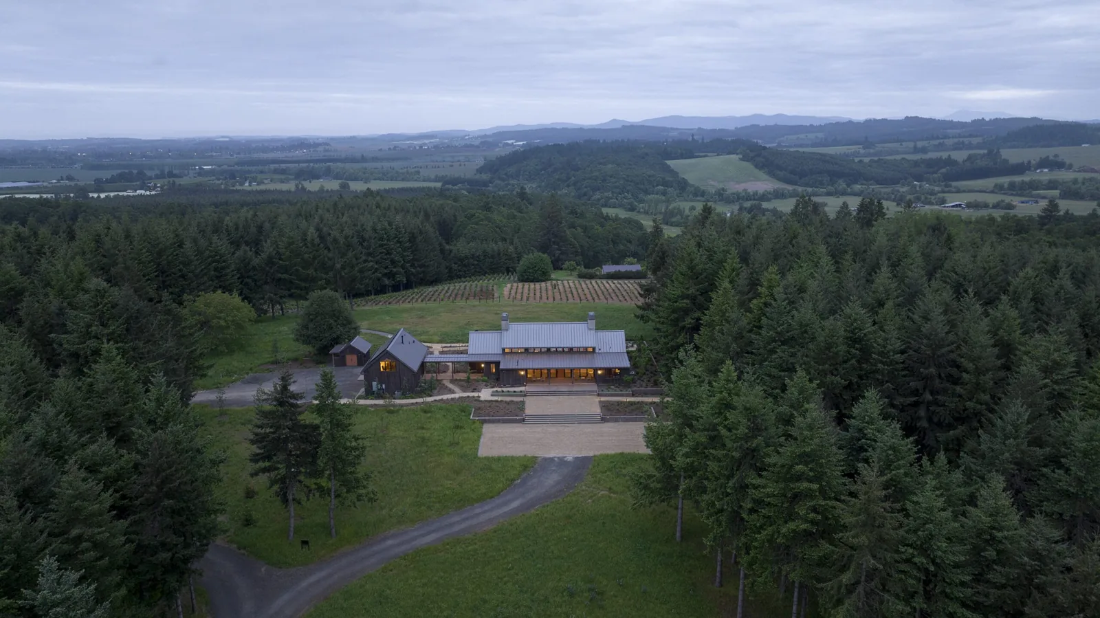 A modern rural building at the edge of dense forest and vineyards shown in an aerial wide view at dusk.
