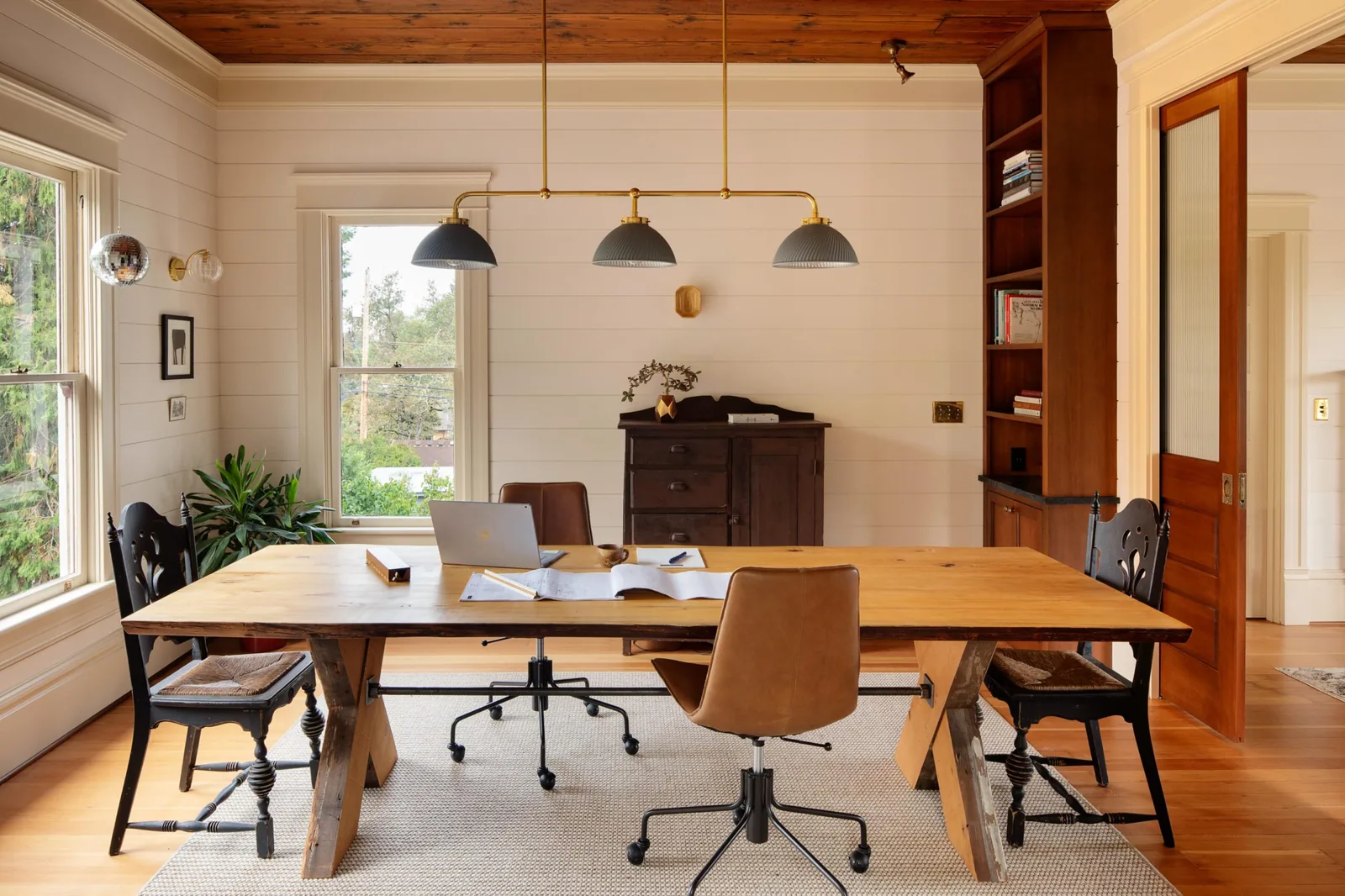 Work area with wooden table, chairs, shiplap walls, and wood ceiling in a mid-size room with natural light from double-hung windows.