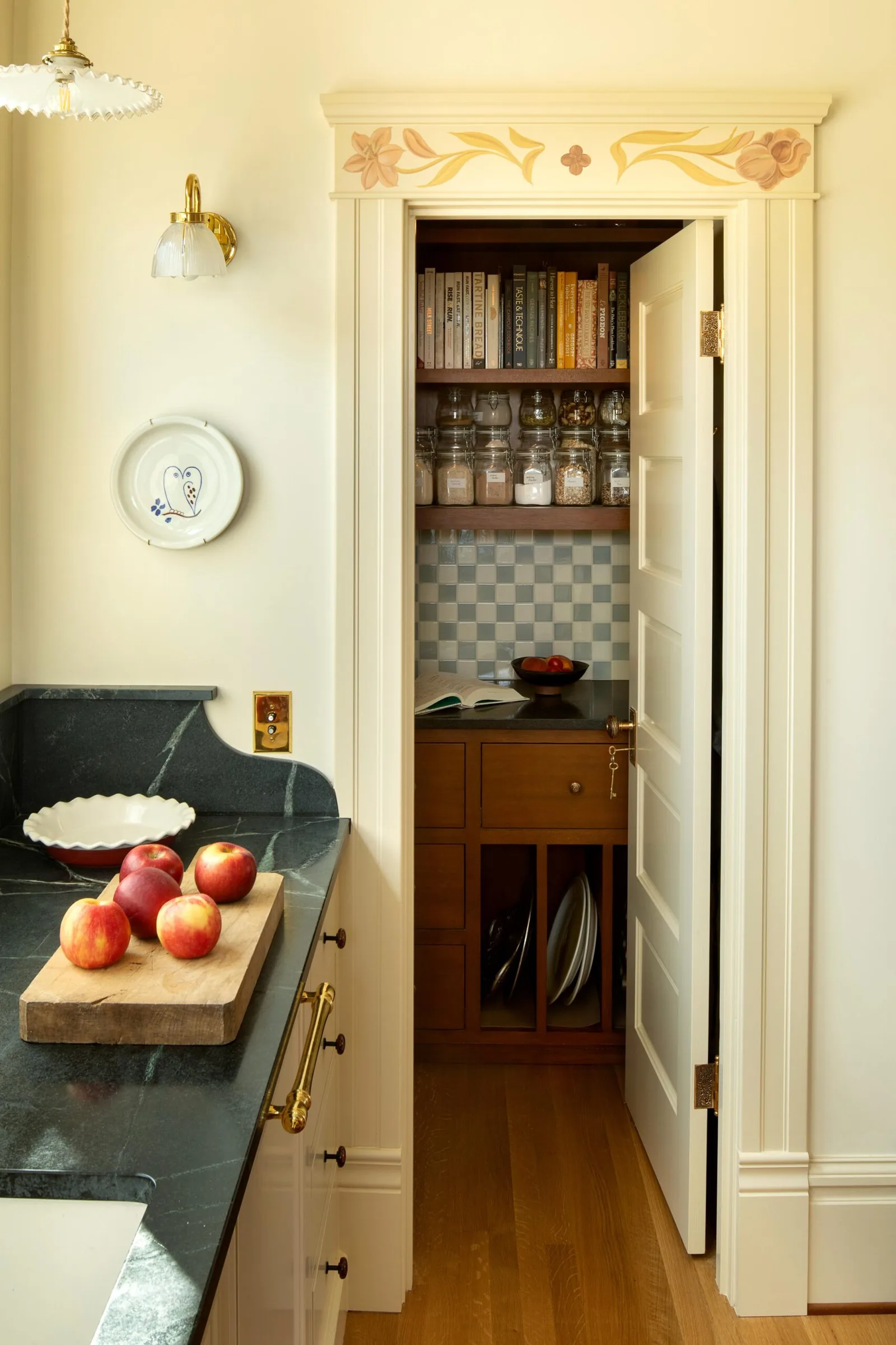 Kitchen with dark stone counter, brass hardware, and adjoining pantry with wooden shelving and tiled backsplash.