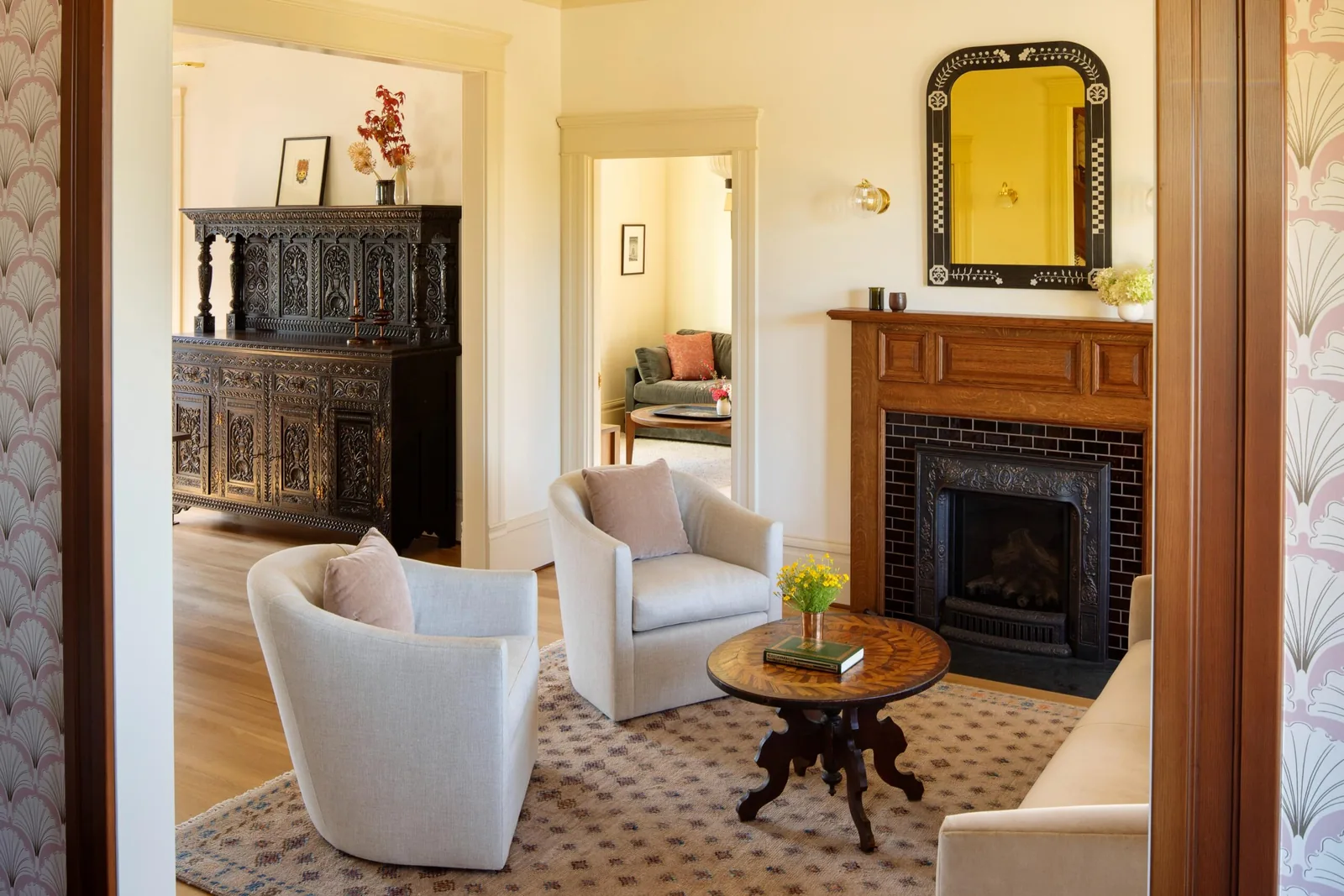 A living room features light upholstered armchairs around a wooden coffee table set on a patterned rug with a wooden fireplace and decorative mirror.