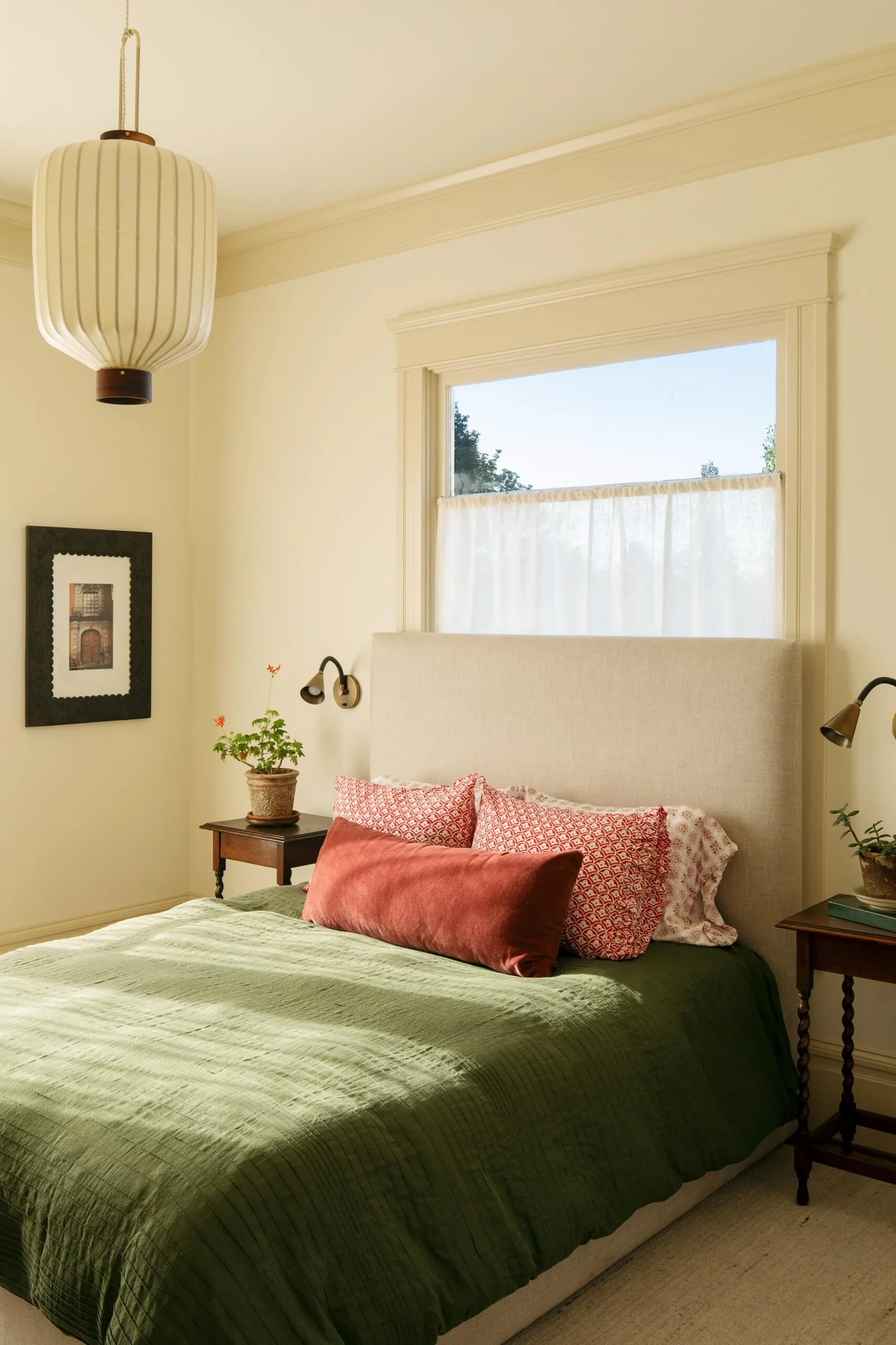 Bedroom featuring a beige upholstered headboard, green bedding, bedside tables with plants, and natural lighting from a window with a sheer curtain.