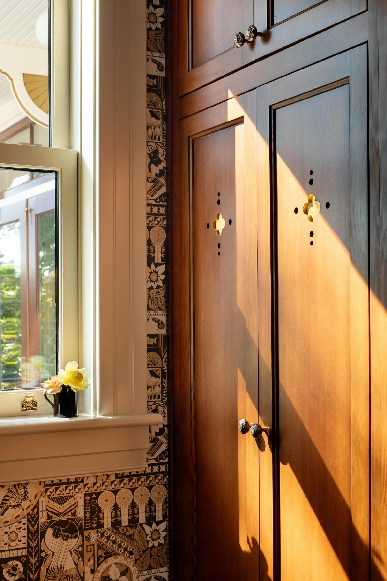 Sunlight illuminates wooden cabinet doors with brass handles adjacent to a window over patterned tile walls.
