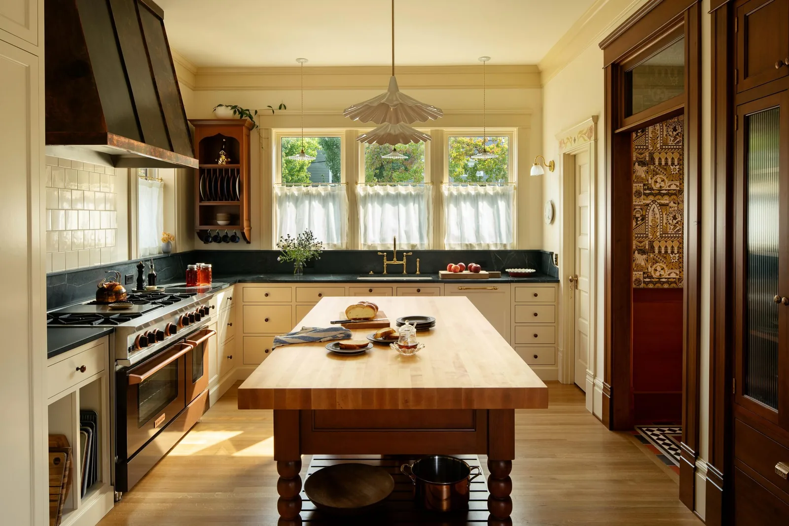 Kitchen with wooden island, black countertops, brass fixtures, and hanging pendant lights in front of three windows.