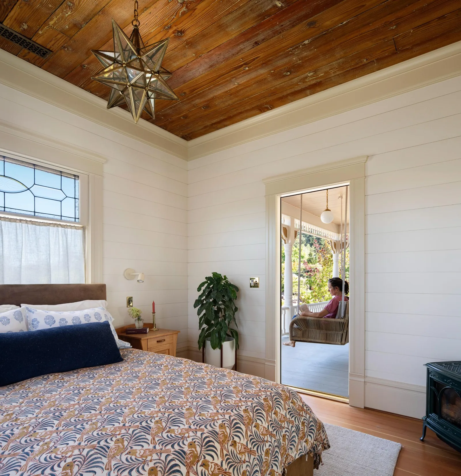 A bedroom features a wooden plank ceiling with a star-shaped pendant light and a door opening to a porch with a hanging chair.