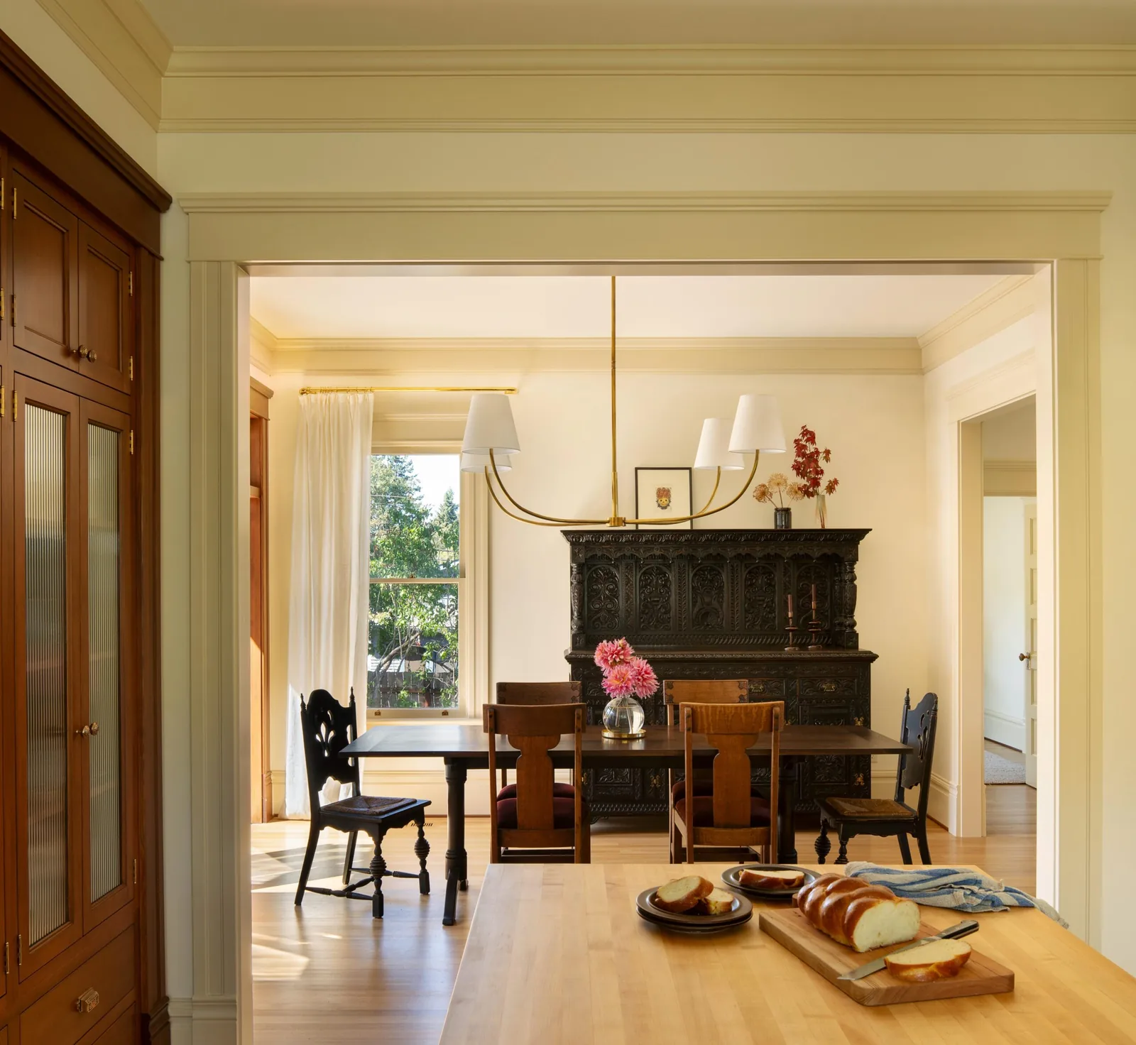 Dining room with traditional carved sideboard, chandelier, wooden chairs, and view from kitchen island with bread on cutting board.