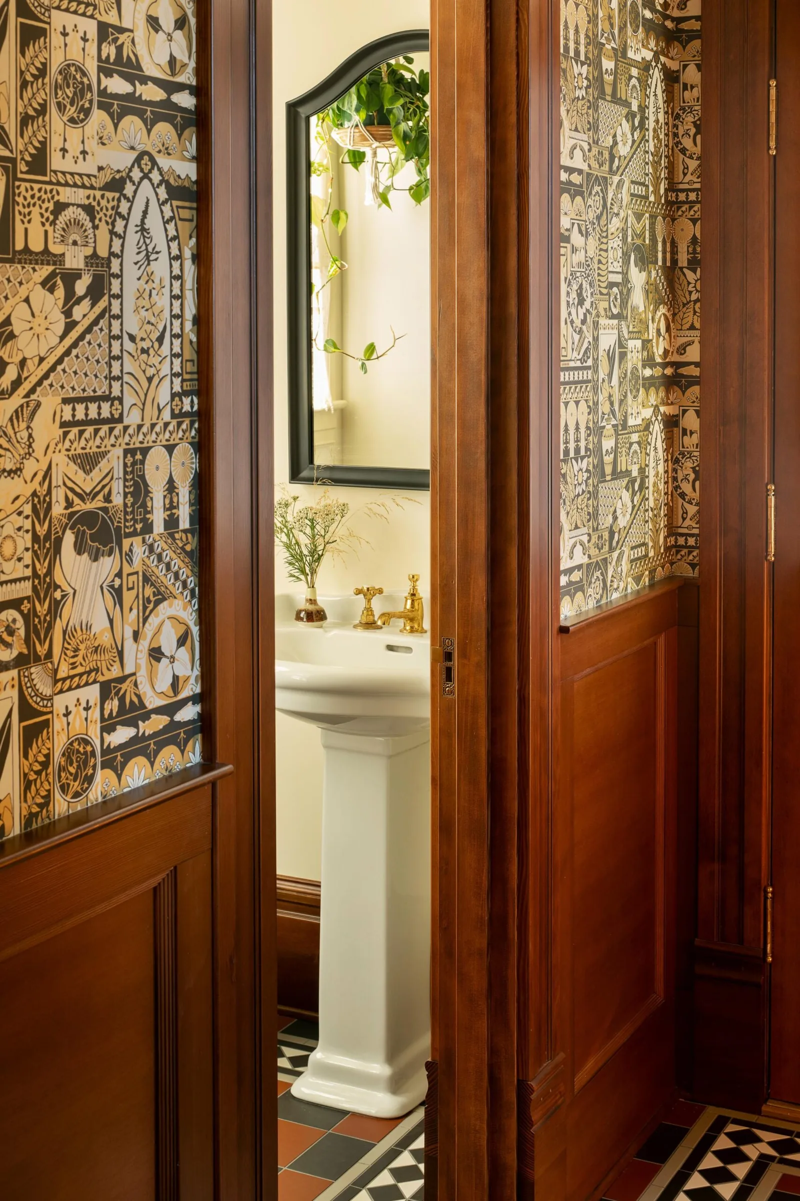 A pedestal sink with brass faucets is visible through a partially open wood-paneled door with patterned wallpaper inside the bathroom.