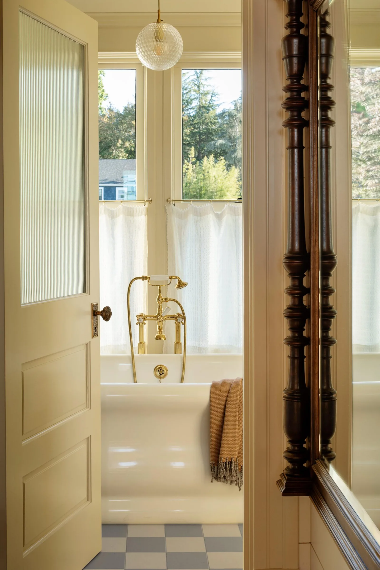 Bathroom with clawfoot bathtub, brass fixtures, checkered floor, and large window with white curtains.