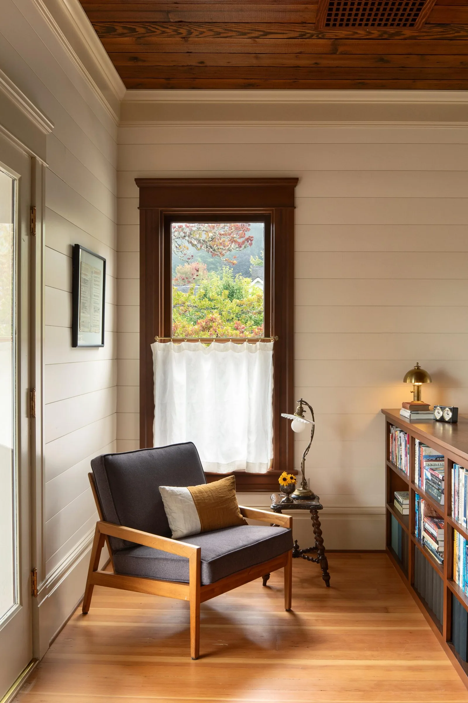 Cozy room corner featuring wooden armchair, bookshelves, and a window with a white curtain.