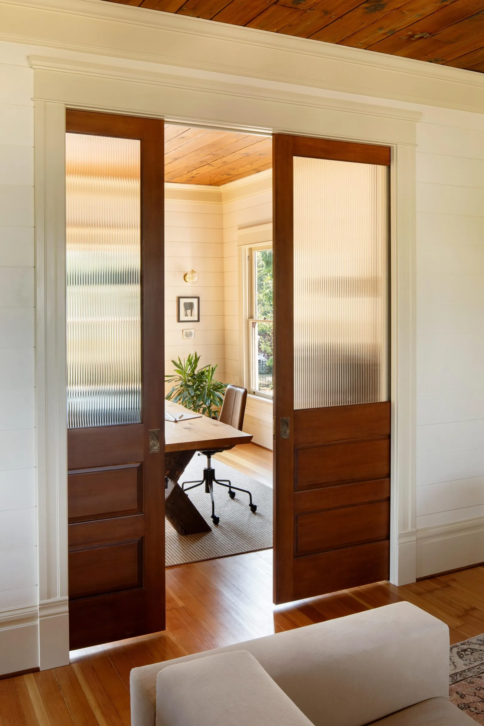 Dark wooden sliding doors with ribbed glass panels separate a living area and an office space with wood flooring.