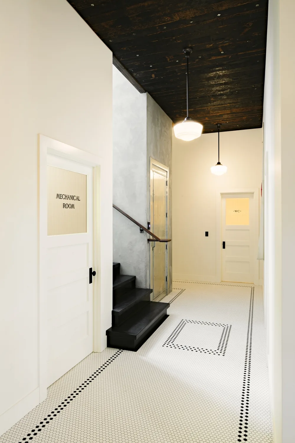 Hallway in the Division Street Residence features white subway-tile walls, black stair railing, and hexagonal tile flooring.