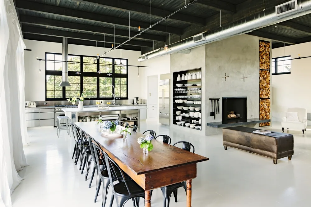 Open kitchen and dining space featuring industrial windows, a concrete fireplace with wood storage, and a wooden dining table.