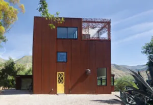 Exterior front view of a rust-colored metal building with varied sized square windows and a yellow door.