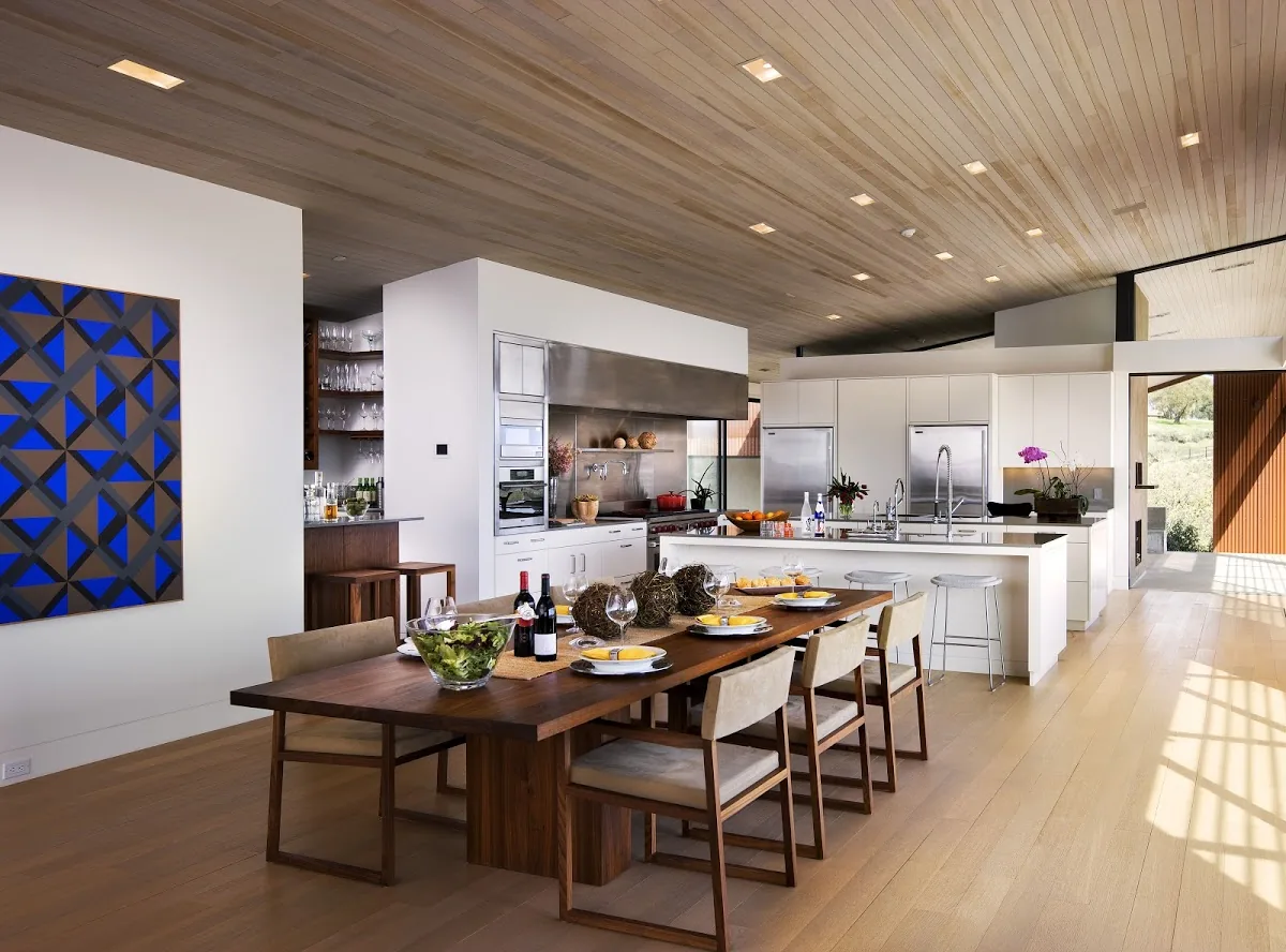 The Santa Ynez Residence kitchen features white cabinetry, wood ceiling, and a large dining table set for a meal.
