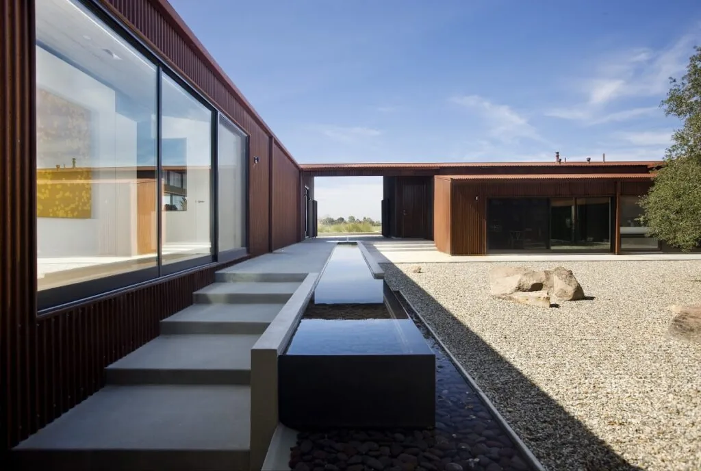 Santa Ynez Residence features a courtyard with a long reflective water element and gravel landscaping, highlighting its rust-colored siding.