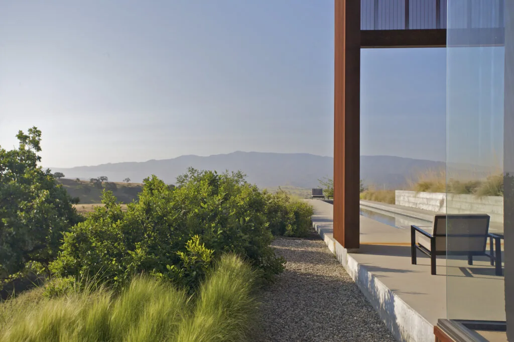 Santa Ynez Residence features a rust-colored exterior with glass walls and a gravel landscape overlooking hills.
