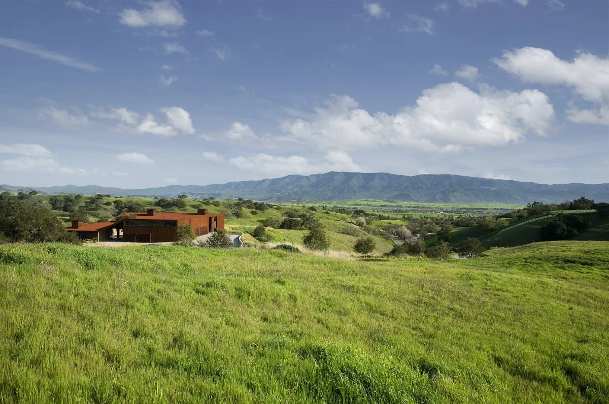 The Santa Ynez Residence by Frederick Fisher and Partners showcases a rust-colored modern home amid expansive green hills and mountain views.