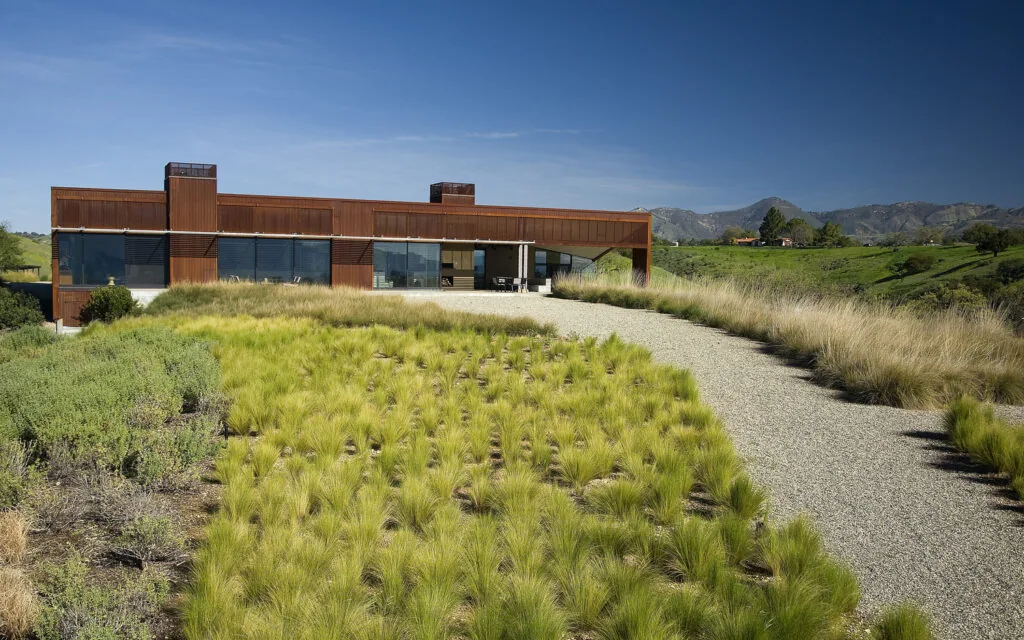 Santa Ynez Residence shows rust-colored metal siding on a single-story home with extensive windows and gravel driveway in a natural hillside setting.