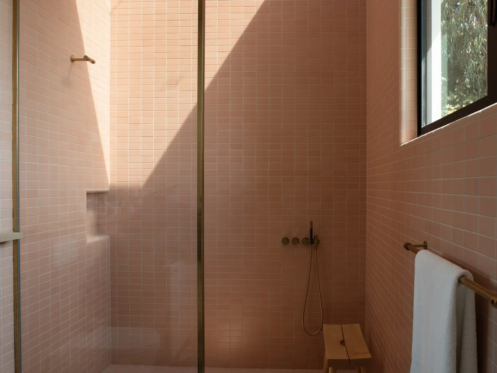 Shower area tiled in pink with brass shower fixtures, a wooden bench, and a towel rack beneath a window.
