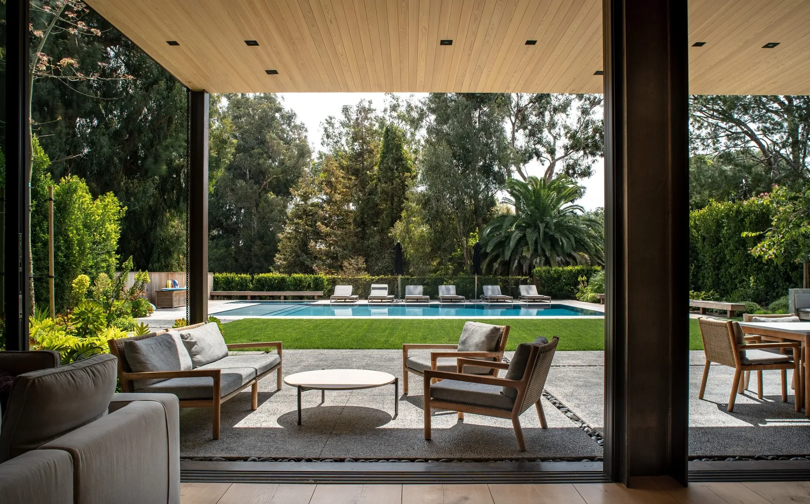 Covered patio with wooden ceiling and seating area opening to a rectangular swimming pool and landscaped garden.