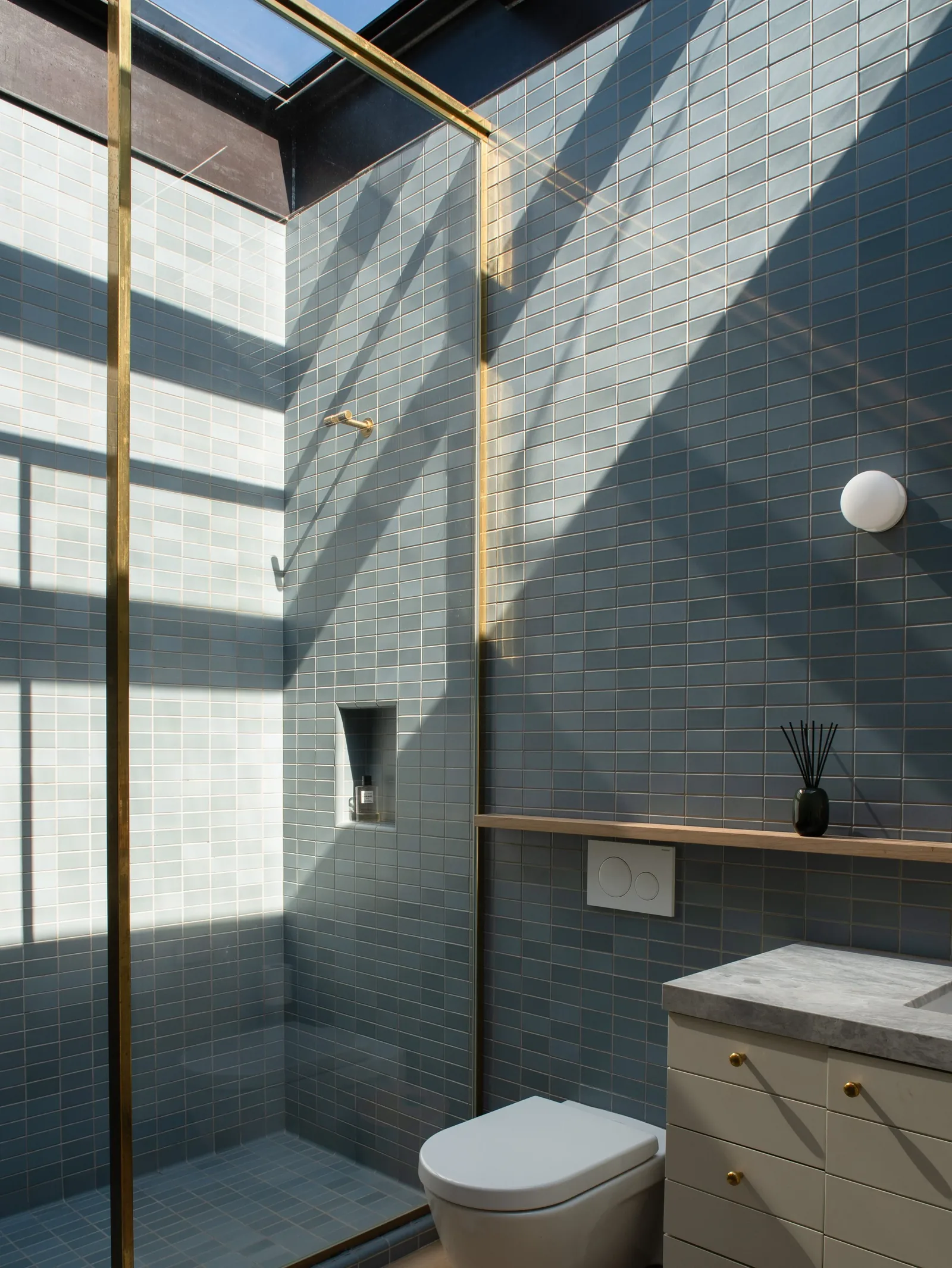 Bathroom with walk-in shower enclosed by brass framing and a skylight above, blue-gray tiled walls and floor, a white wall-mounted toilet, and a vanity with a marble countertop.