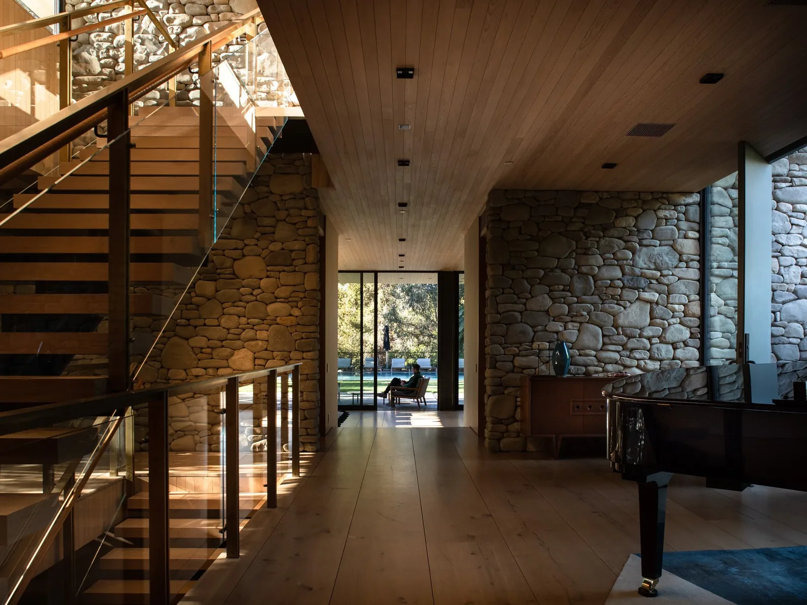 Hallway interior with stone accent walls, wooden ceiling, floating wood staircase with glass railing, and a black grand piano.