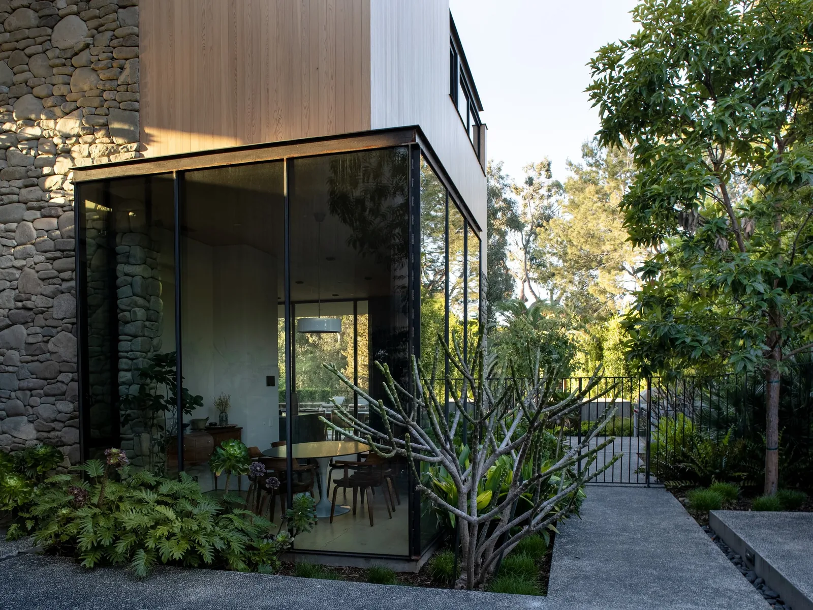 Modern house corner with stone facade, floor-to-ceiling glazing, and wood siding adjacent to a garden pathway.