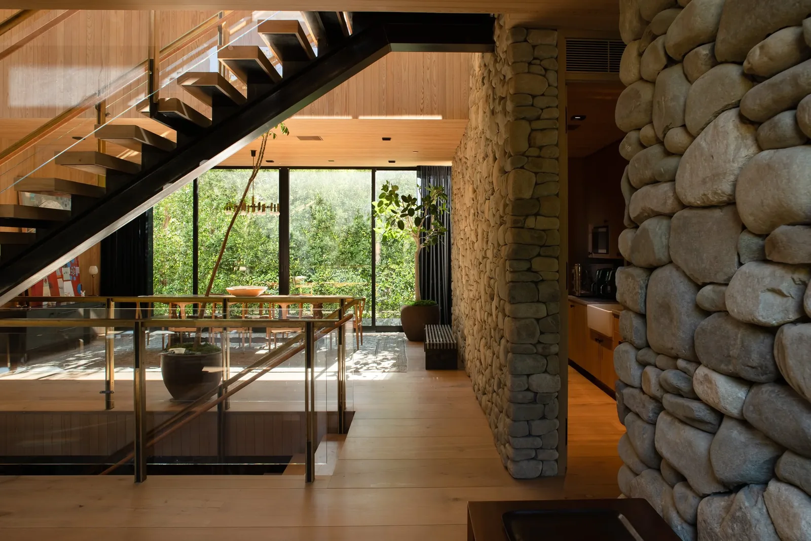 Interior view showing a floating staircase with wood treads and floor-to-ceiling glazing behind a dining area with stone accent walls.