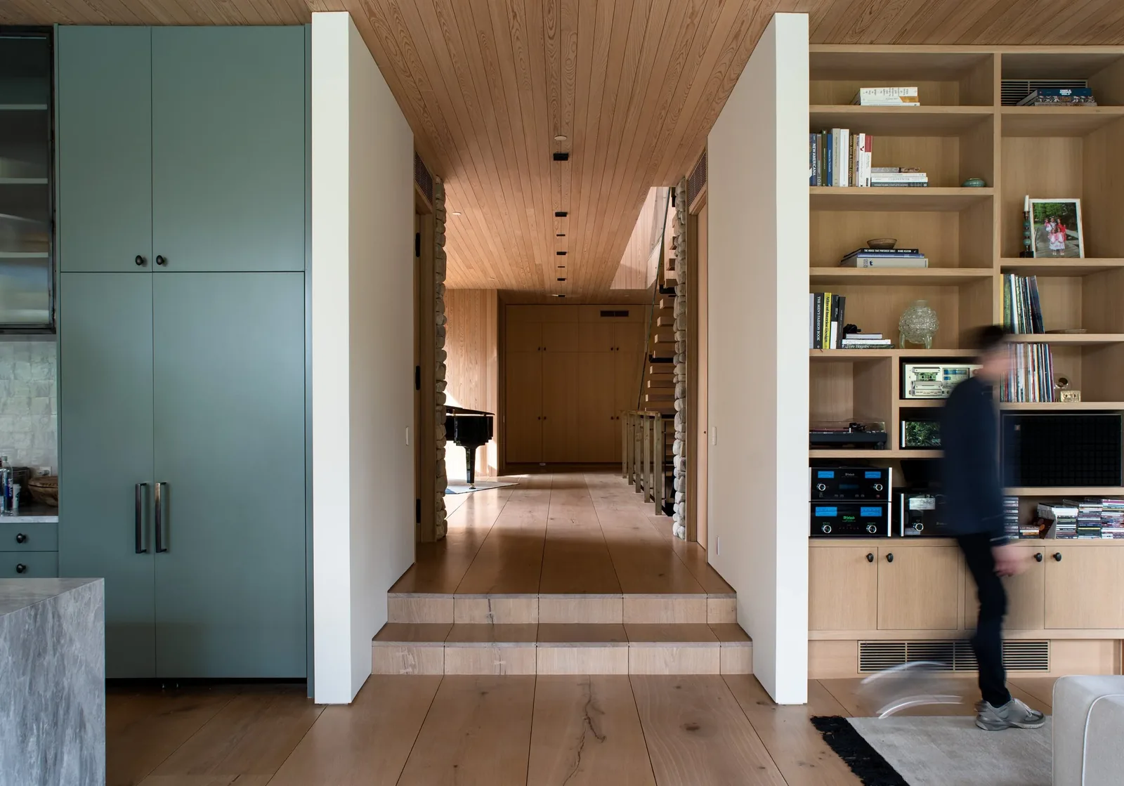 Hallway with wooden flooring and ceiling, adjacent to a kitchen area with green cabinetry and built-in shelving unit on the right.
