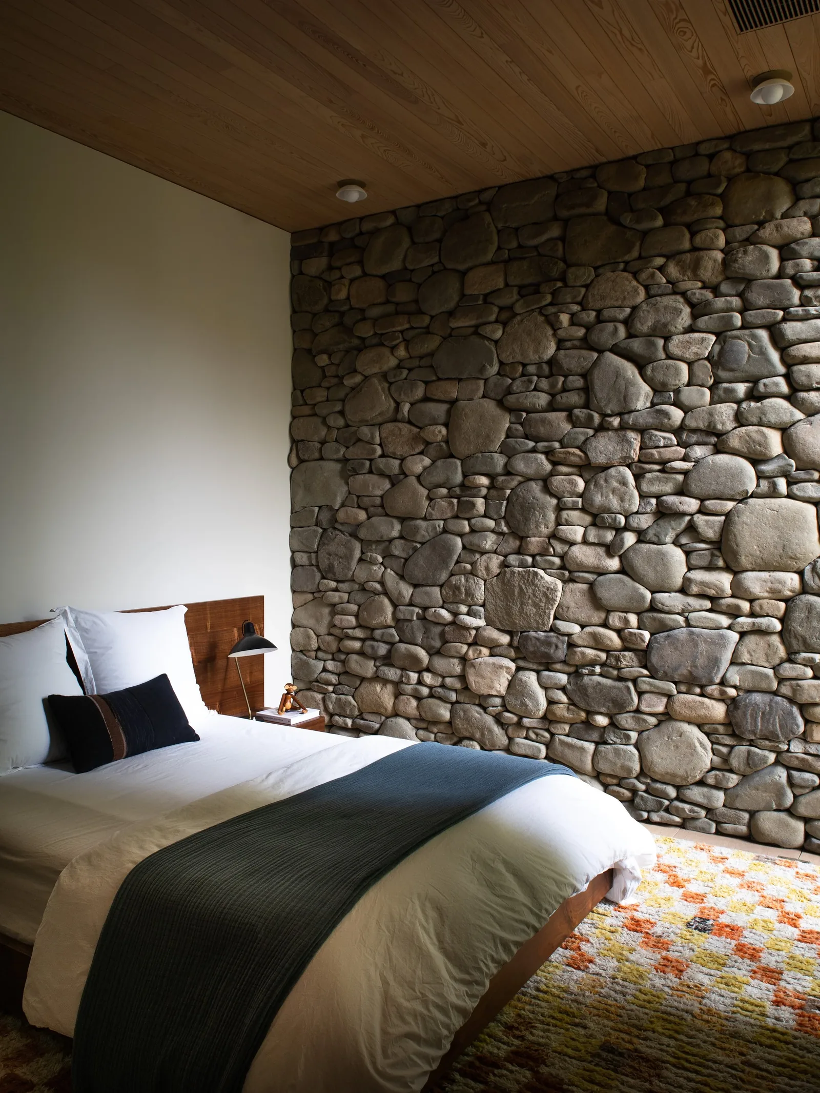 A bedroom features a textured stone wall, wooden ceiling, and simple bedding with a decorative throw.