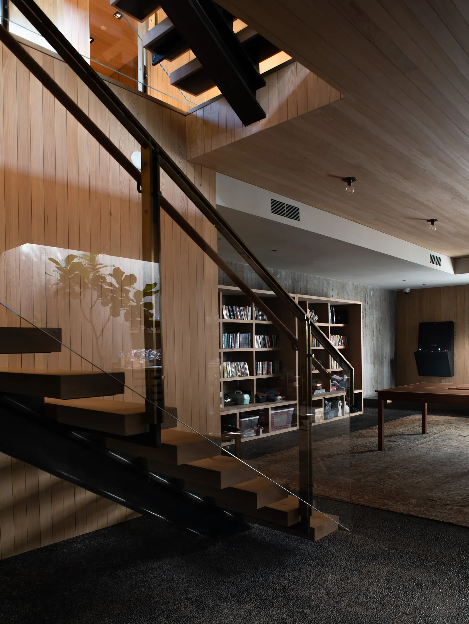 Modern staircase with wood treads and glass railing adjacent to a room with built-in bookshelves and carpet.