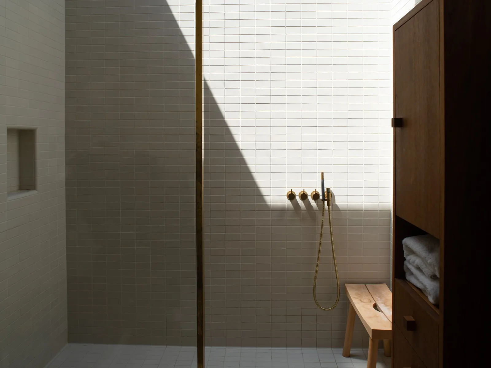 A minimalist shower with white tiled walls and floor, brass shower controls, a wooden bench, and a wooden storage cabinet with towels.